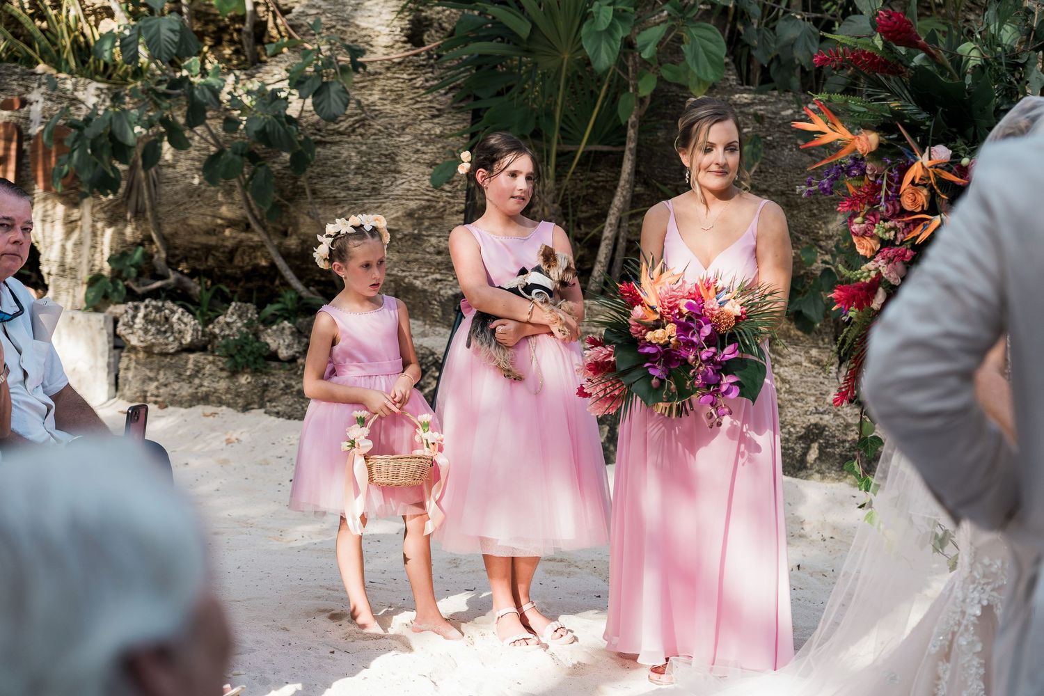 Bridesmaids in pink dresses holding tropical bouquets stand with flower girls at an outdoor beach wedding ceremony.