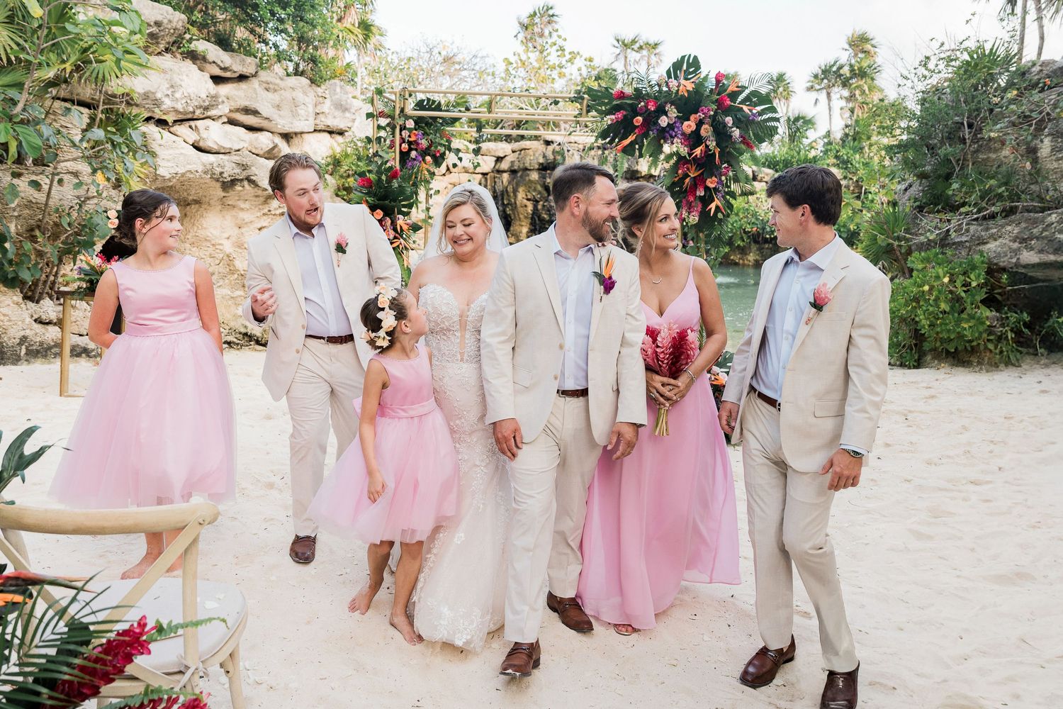 Wedding party walks on sandy beach in pink and beige attire with tropical floral arch backdrop.