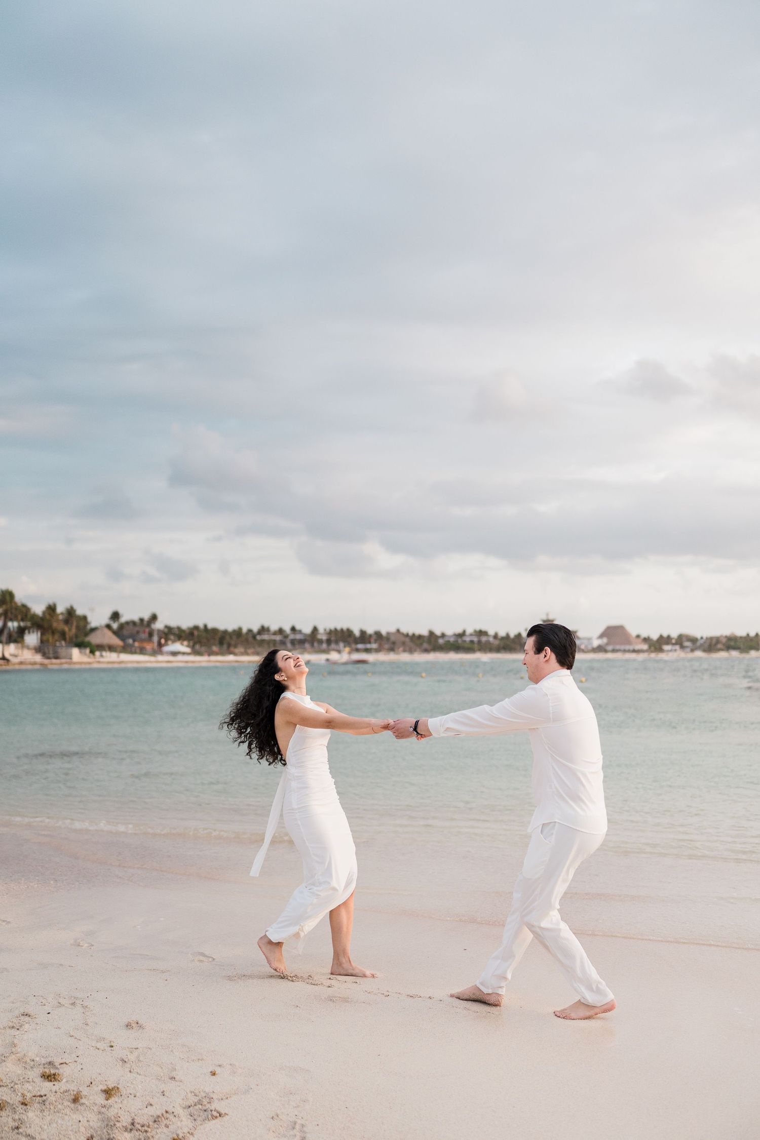 Couple in white outfits holding hands and dancing on a tropical beach with turquoise water in the background.