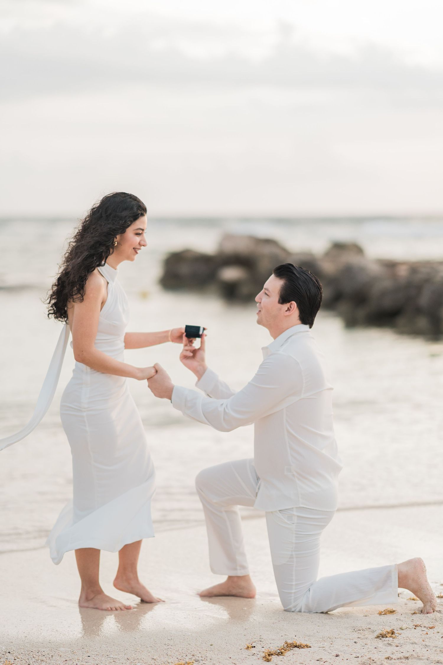 Man proposes to woman on one knee at beach, holding ring box, both dressed in white.