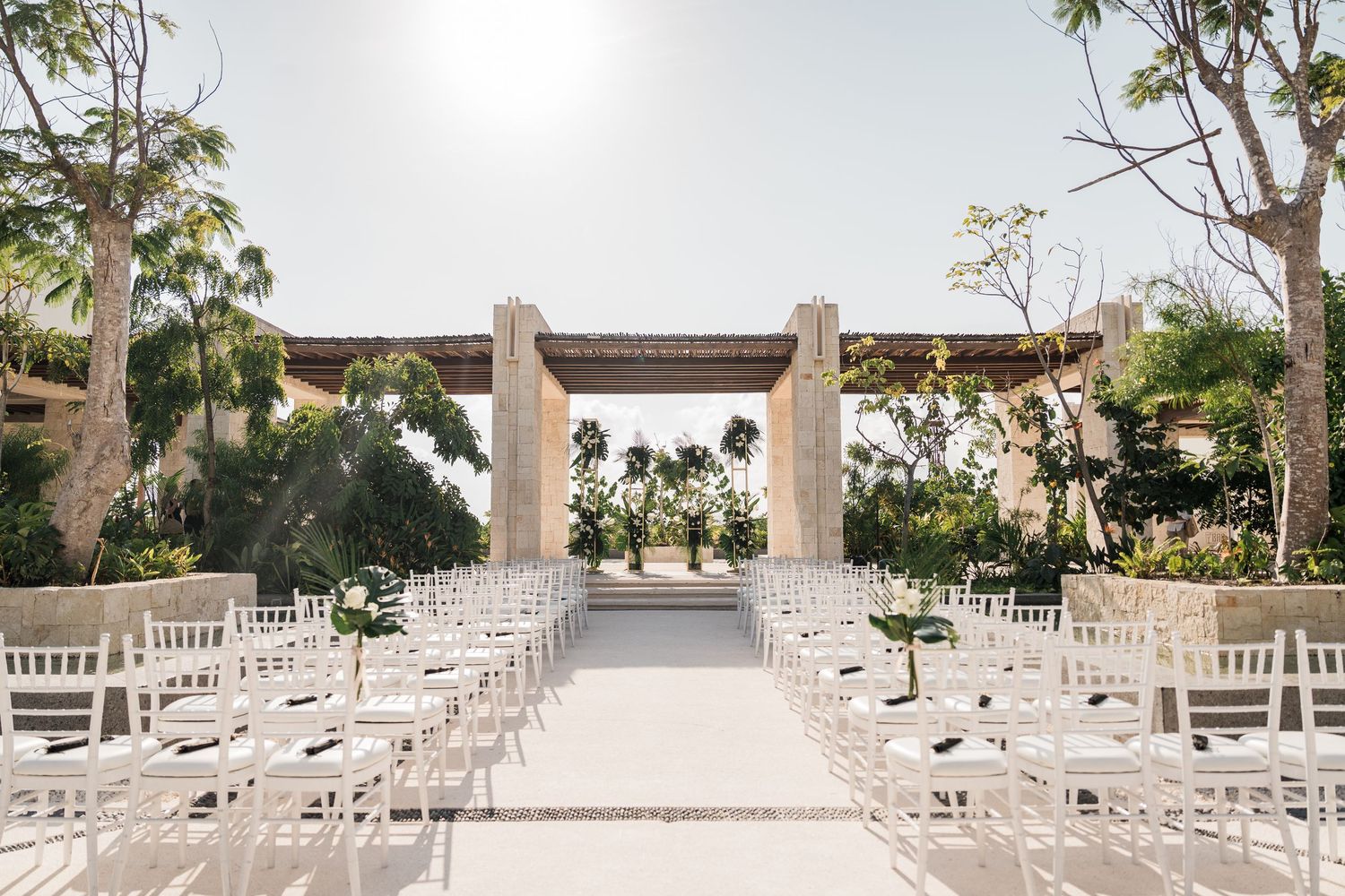 Elegant outdoor wedding ceremony setup with white chairs, floral arrangements, and a stone arch altar surrounded by tropical trees.