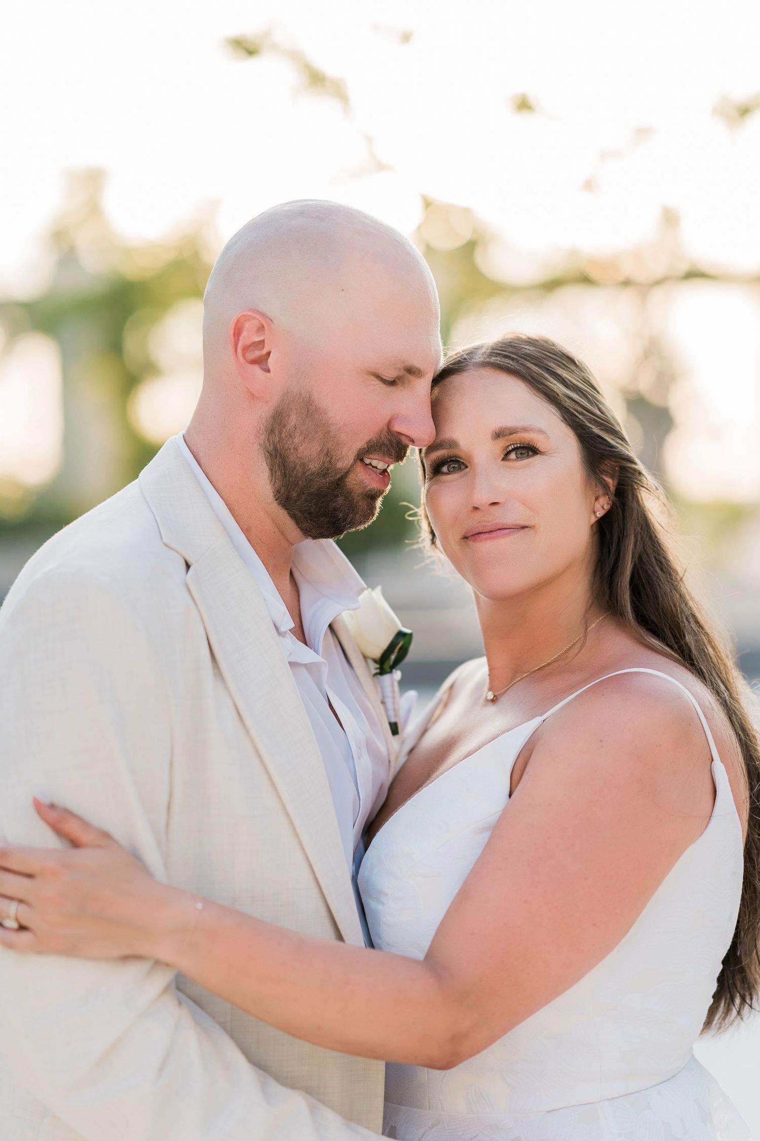 Bride and groom share a romantic golden hour moment, she in white gown, he in tan suit with white boutonniere.