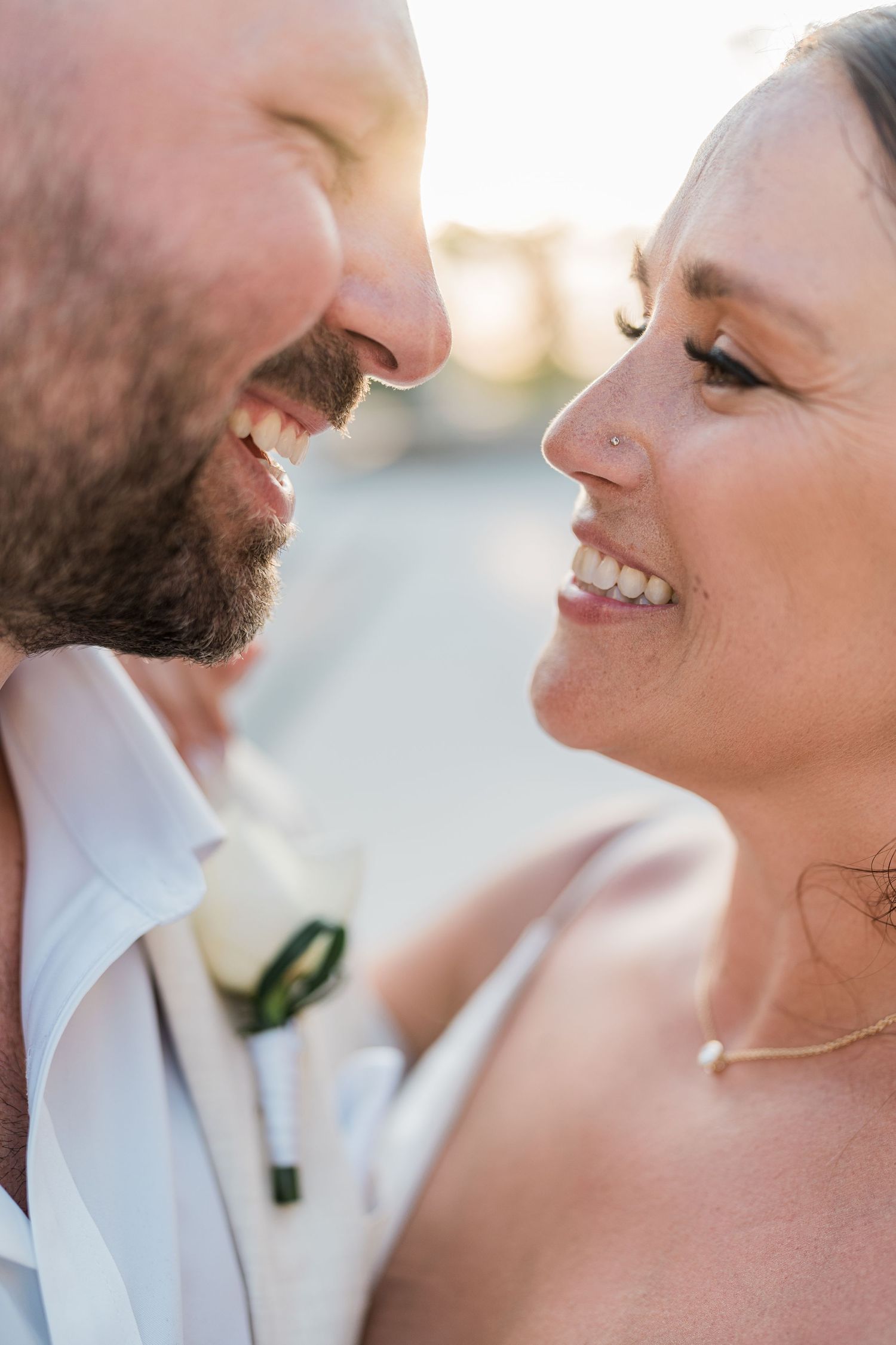 Close-up of smiling bride and groom facing each other at golden hour, groom wearing white shirt with boutonniere.