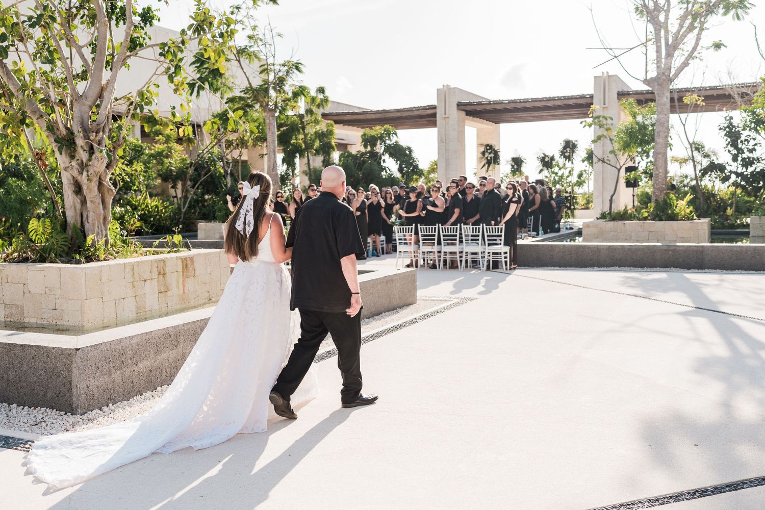 Bride and groom walking toward outdoor wedding ceremony with guests seated in white chairs under a modern pergola.