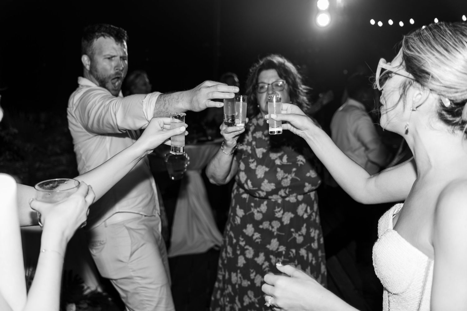 Black and white photo of wedding guests toasting with drinks on the dance floor at a festive reception.