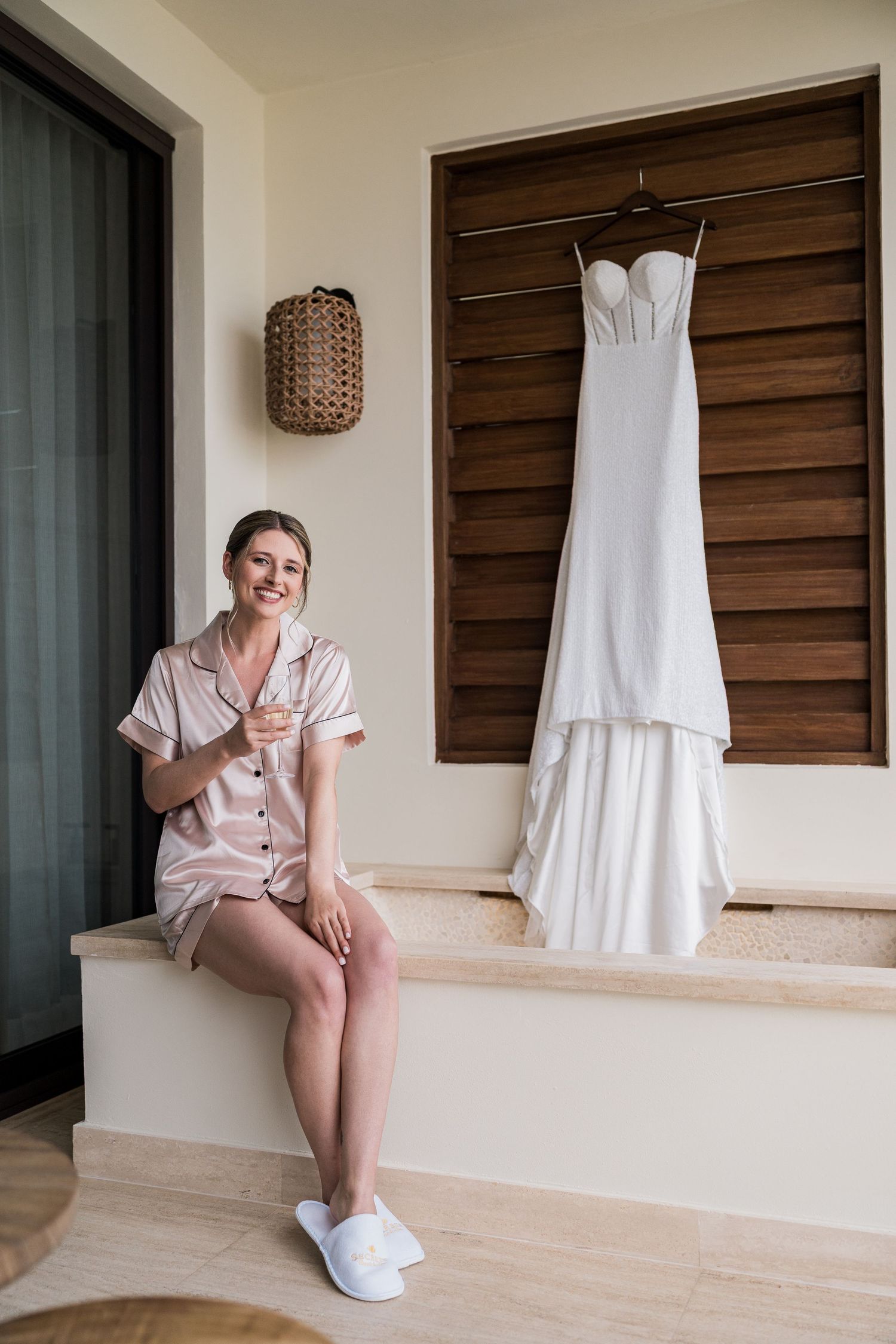 Smiling bride in pink satin pajamas holding champagne beside her white wedding gown hanging on wooden shutters.