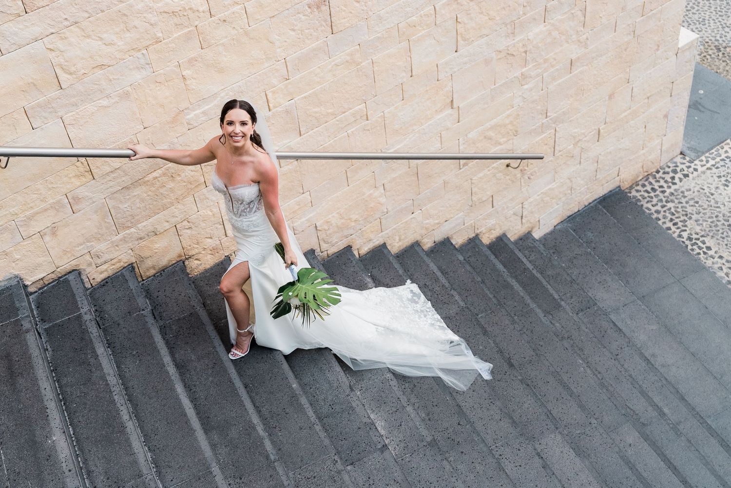 Bride in white gown holding bouquet poses on outdoor stone staircase, smiling elegantly on her wedding day.