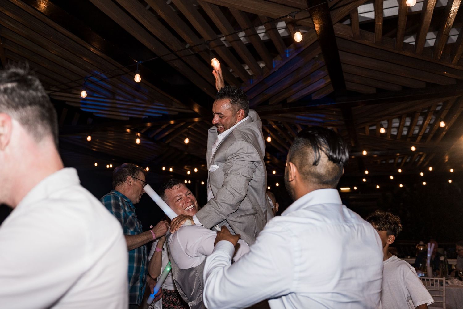 Groom lifted by groomsmen on dance floor at rustic wedding reception with string lights.