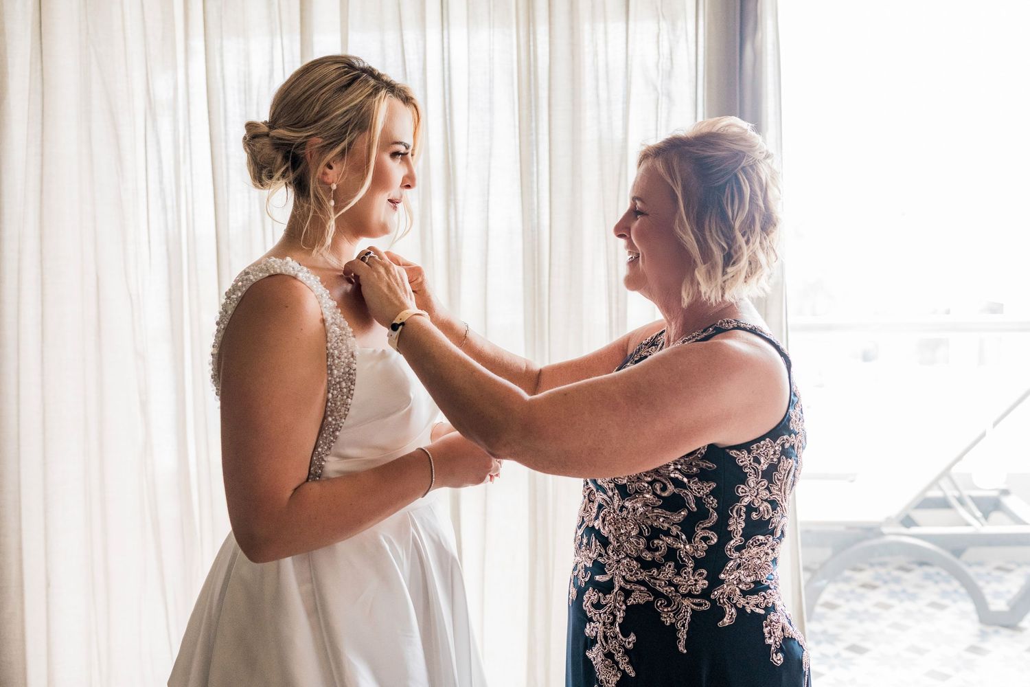 Bride in white gown gets necklace fastened by smiling woman in embroidered dress near sheer curtains.
