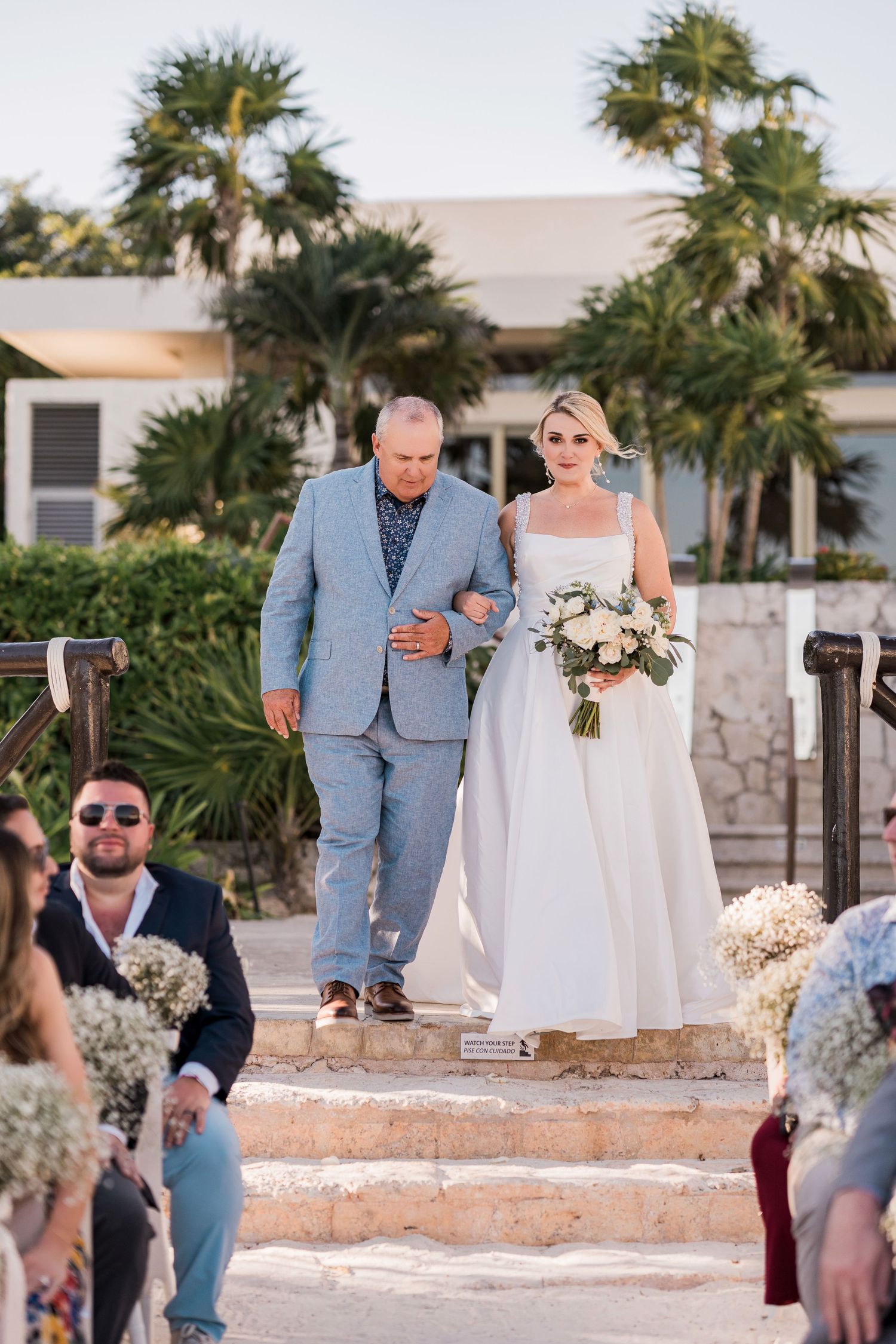 Bride in white gown walks down outdoor aisle with escort, holding bouquet, guests seated, palm trees in background.