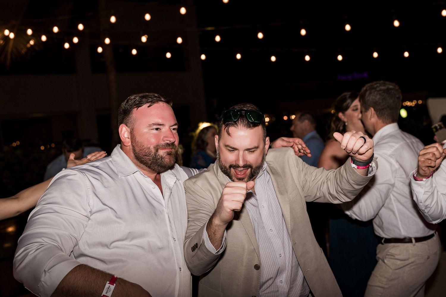 Two men dancing energetically at an outdoor wedding reception under string lights at night.