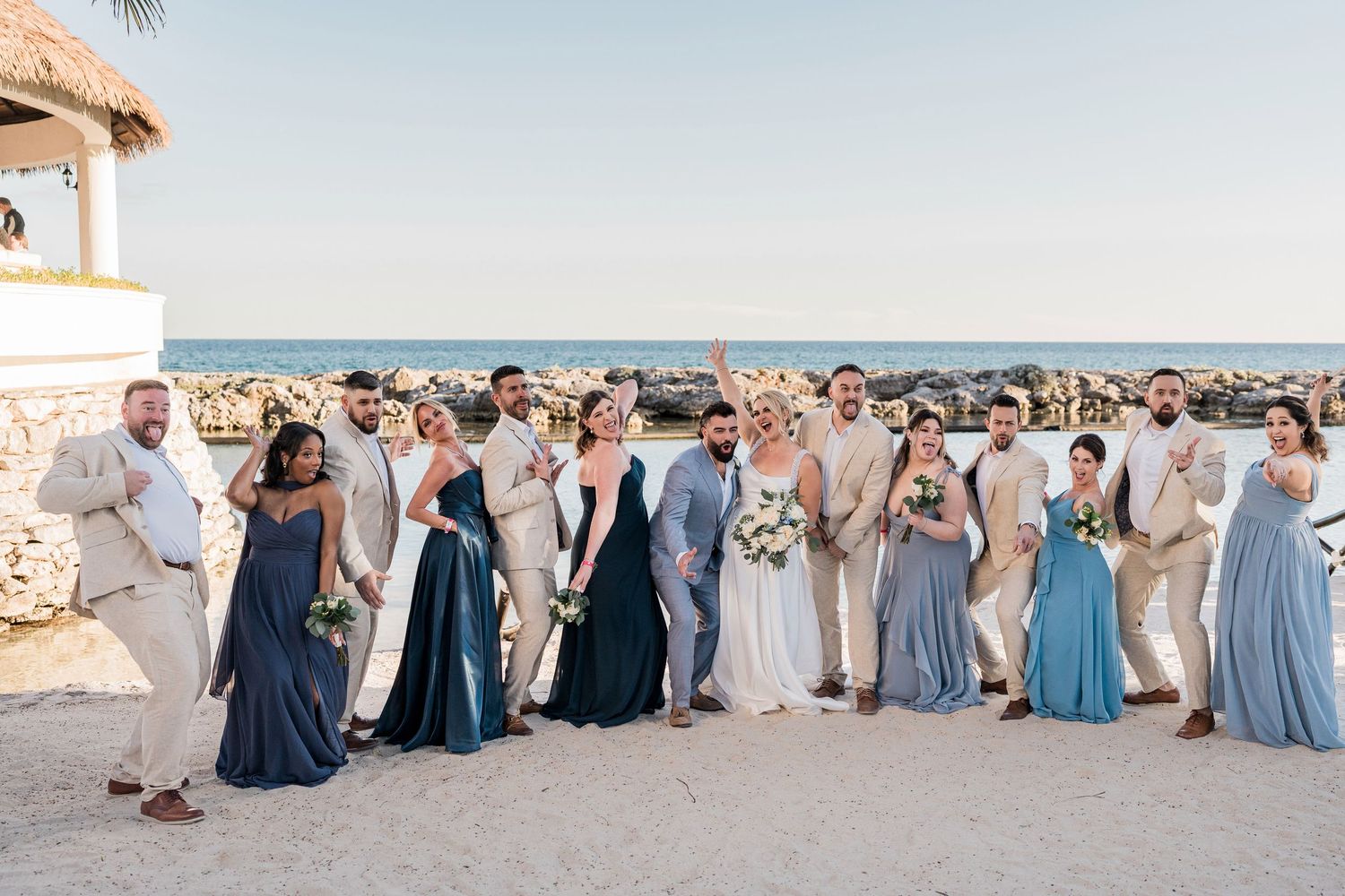 Wedding party poses on a sandy beach near the ocean, bridesmaids in blue gowns and groomsmen in tan suits.