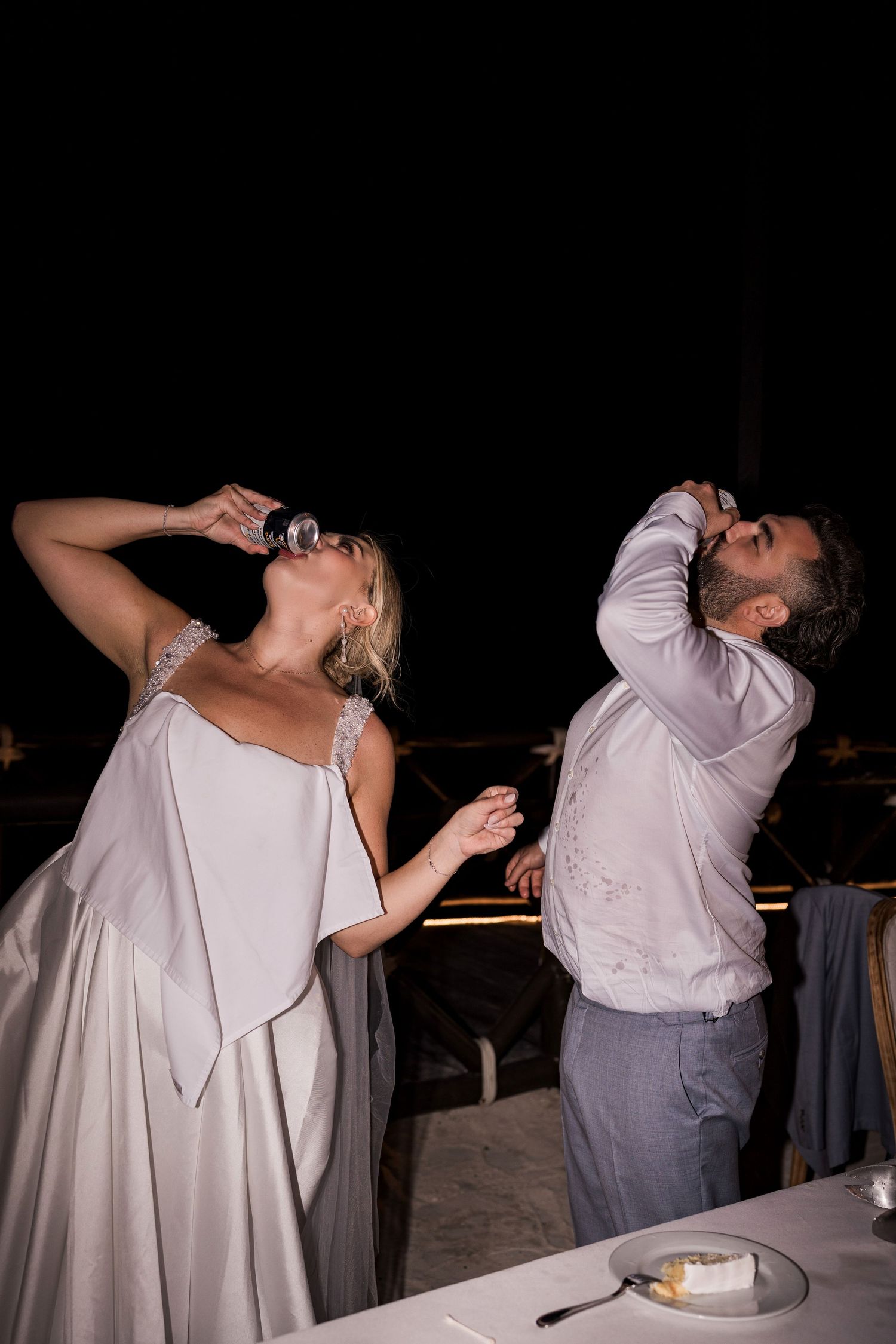 Bride and groom joyfully drinking at their wedding reception outdoors at night, woman in elegant white gown.