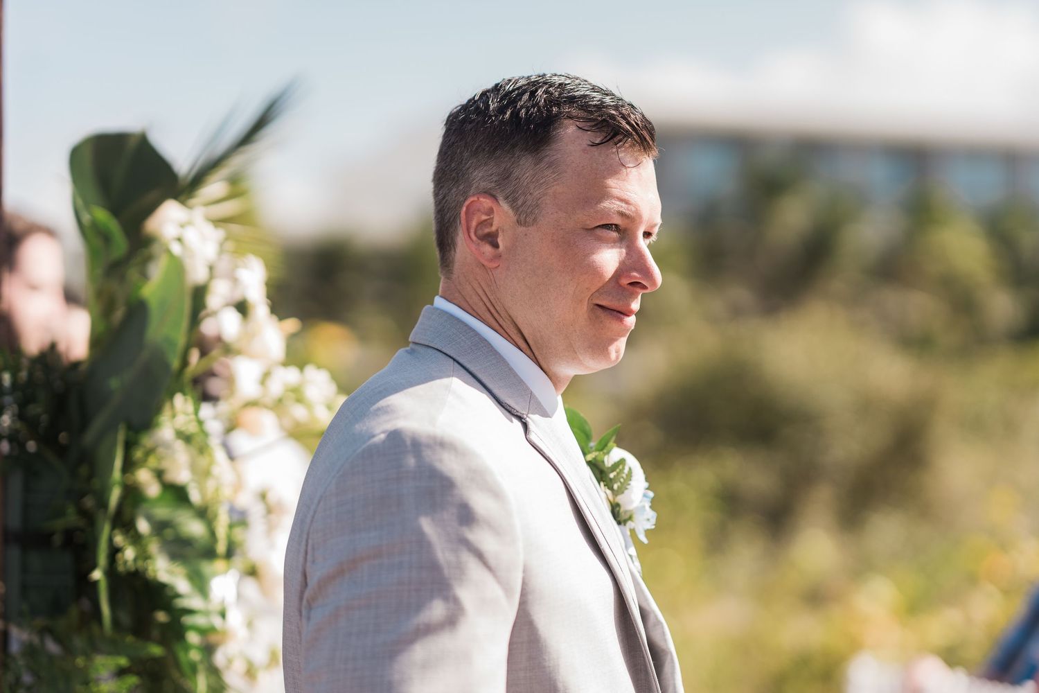 Groom in light gray suit with boutonniere smiling at outdoor wedding ceremony with floral arch backdrop.