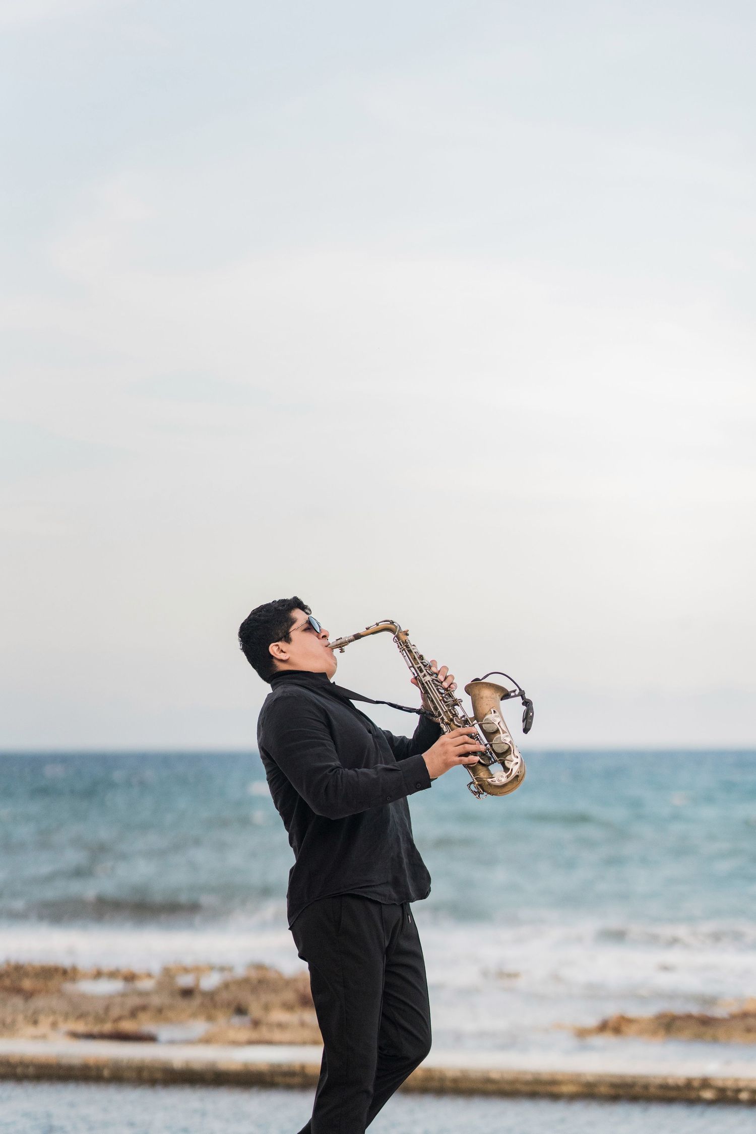 Musician in black suit playing saxophone on rocky beach with ocean waves and cloudy sky in background.