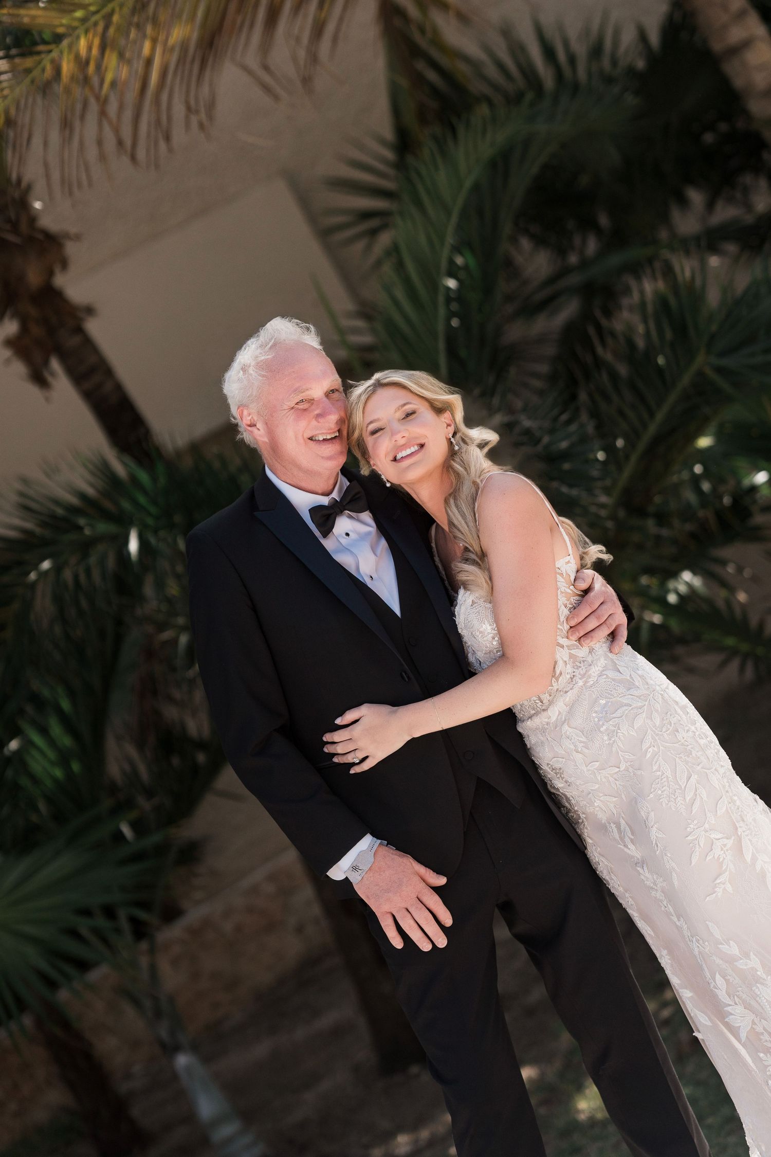 A smiling bride in a lace gown embraces an older man in a tuxedo outdoors with tropical palm trees behind them.