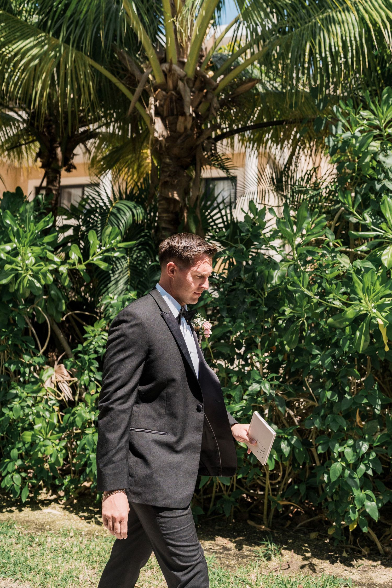 Man in dark suit holding tablet walking outdoors past tropical palm trees and lush green plants on sunny day.
