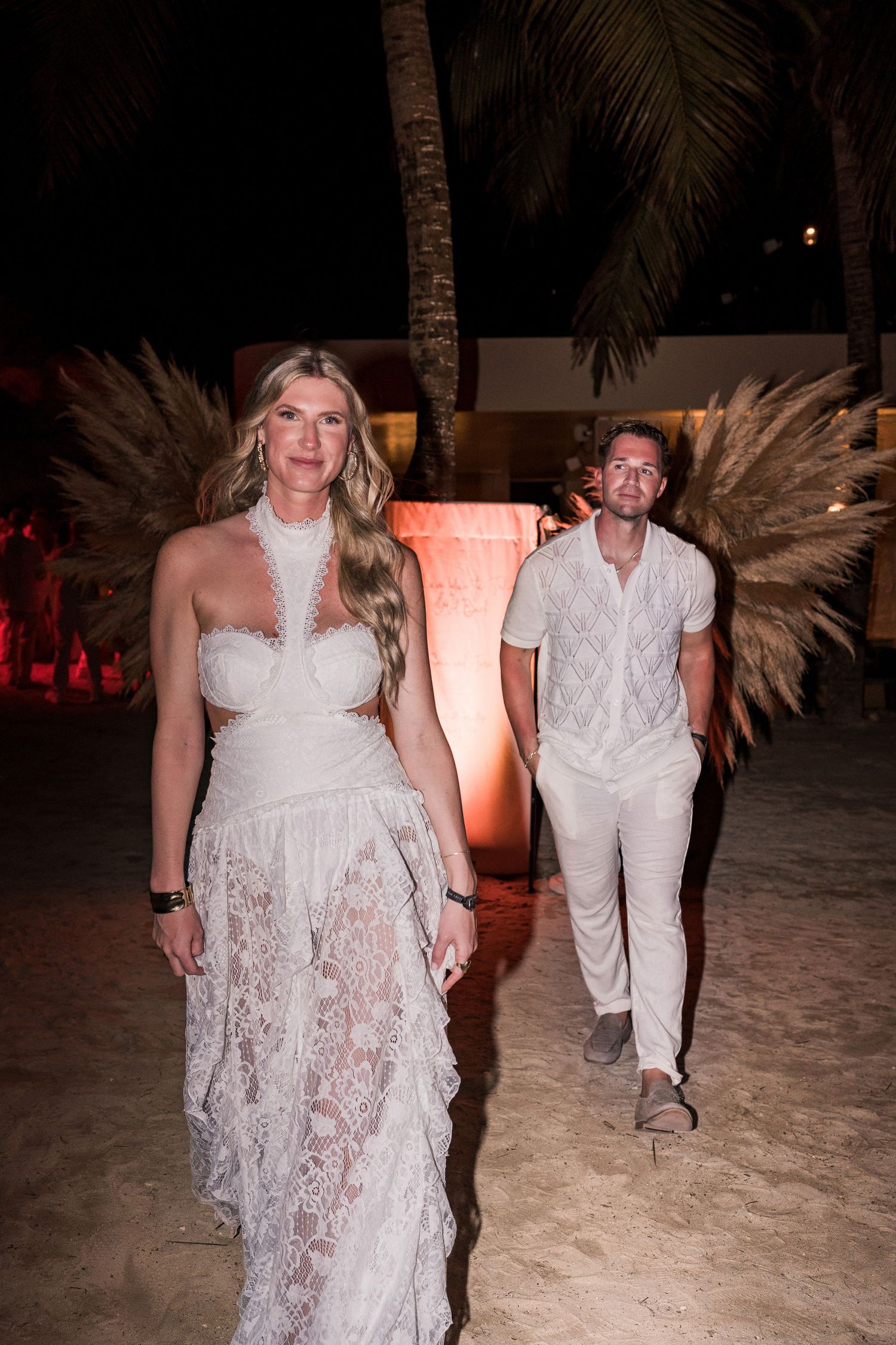 Couple in white outfits walking on a beach at night, woman in lace gown, man in casual white attire, palm trees behind.