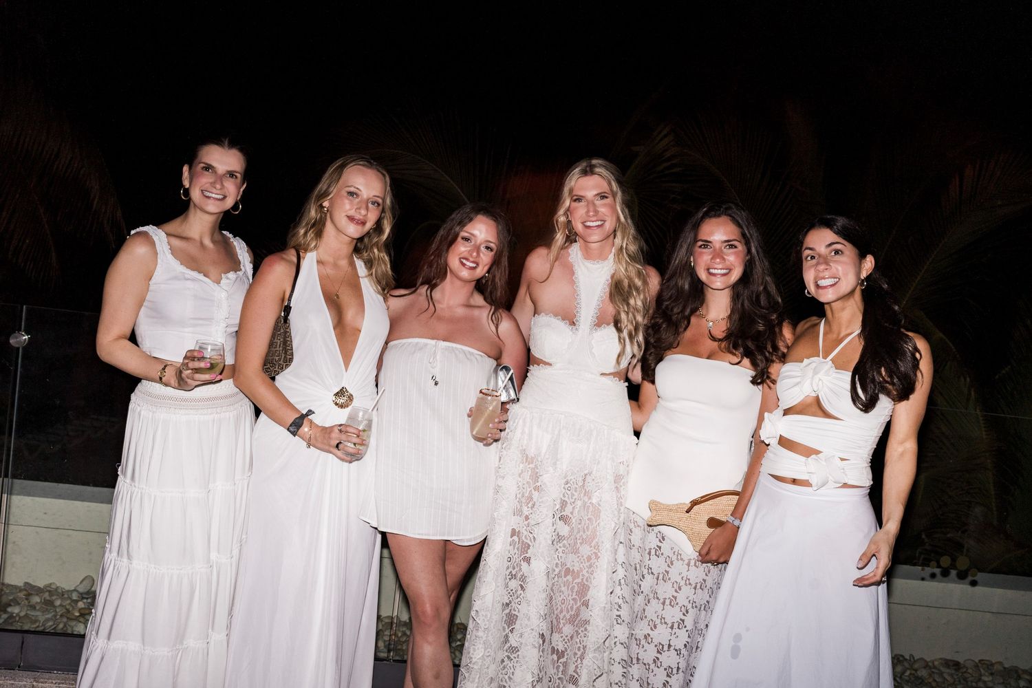 Six women in white outfits smiling together at an outdoor evening event.