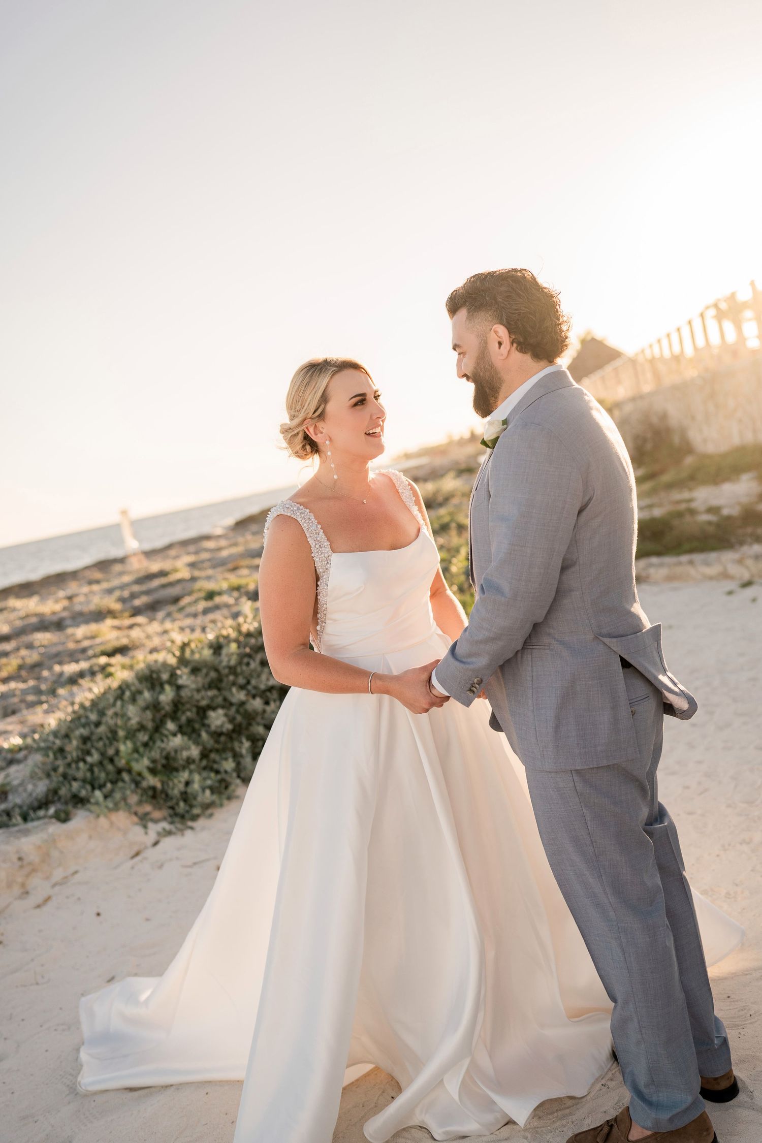 Bride and groom holding hands on a sandy beach at sunset, bride in white gown, groom in gray suit. Puerto Vallarta wedding photographer