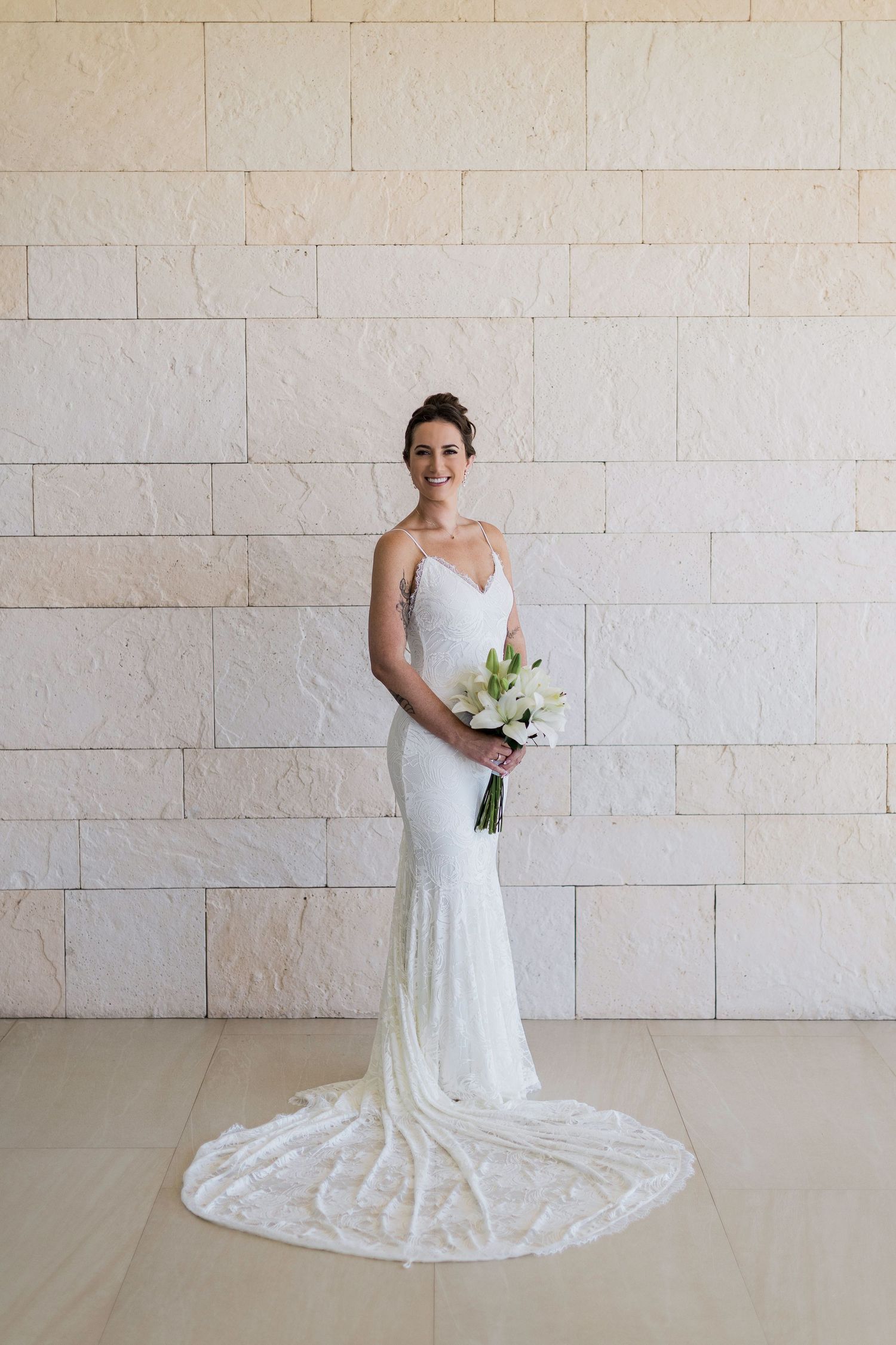 Bride in white fitted spaghetti strap wedding gown with train holds floral bouquet, posing against stone wall.