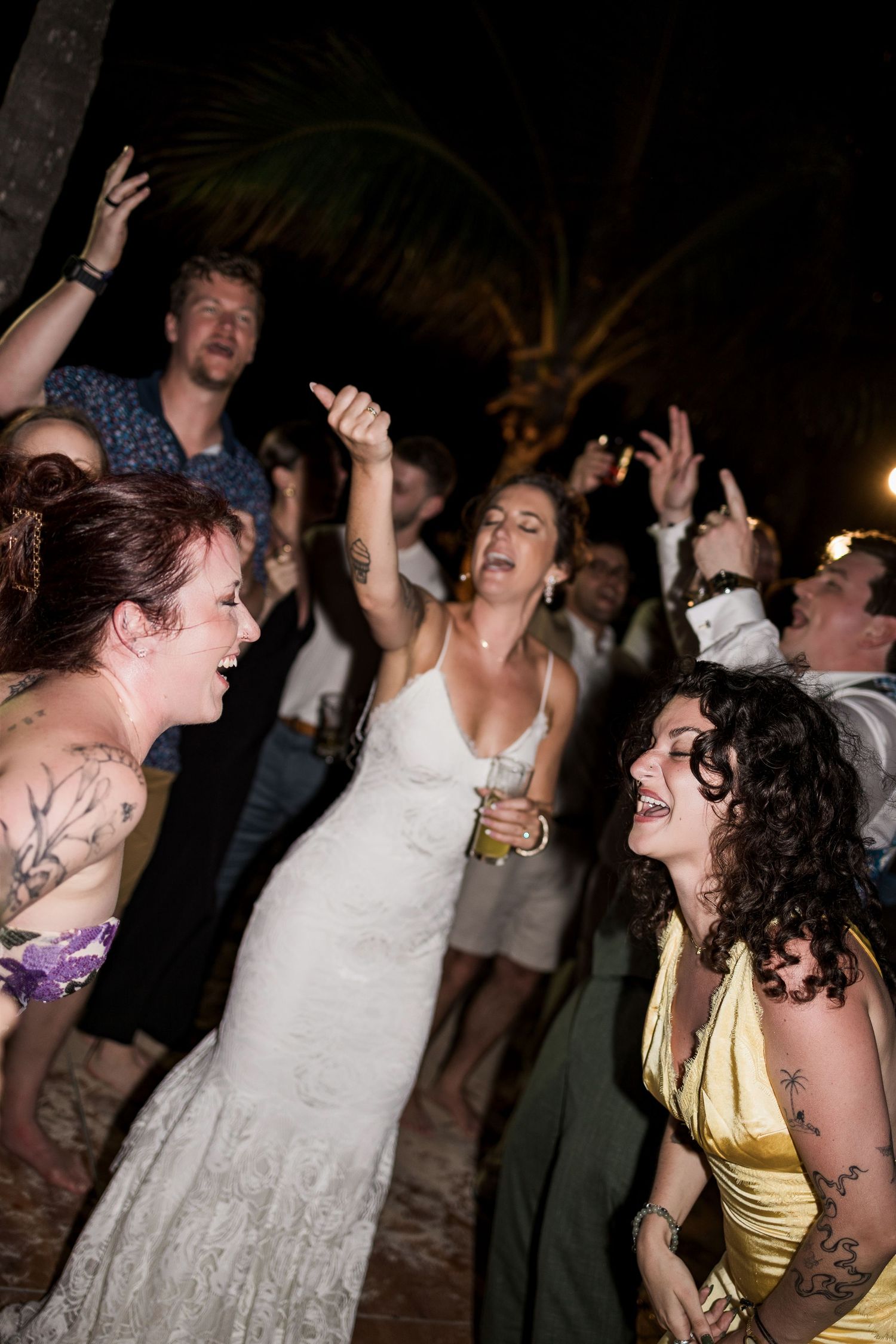 Bride in white dress dancing joyfully with tattooed guests at an outdoor nighttime wedding reception party.