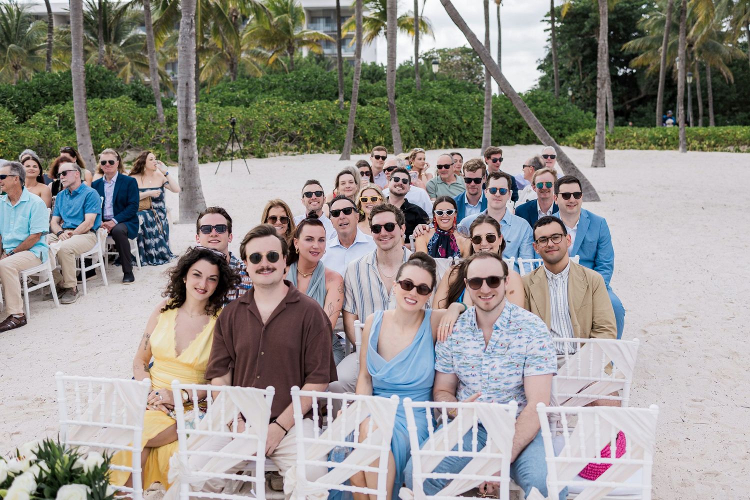 Large group of wedding guests posing together on a tropical beach ceremony with palm trees and white chairs.
