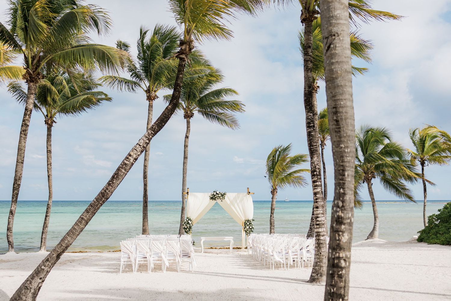 Beach wedding setup with white chairs and floral arch under palm trees on a tropical sandy shore with turquoise ocean views.