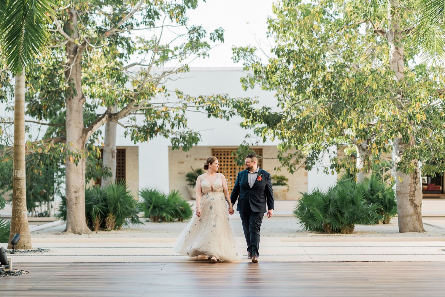 Secrets Akumal Wedding - Bride and groom walk hand-in-hand on a wooden deck surrounded by lush trees outside a white wedding venue.