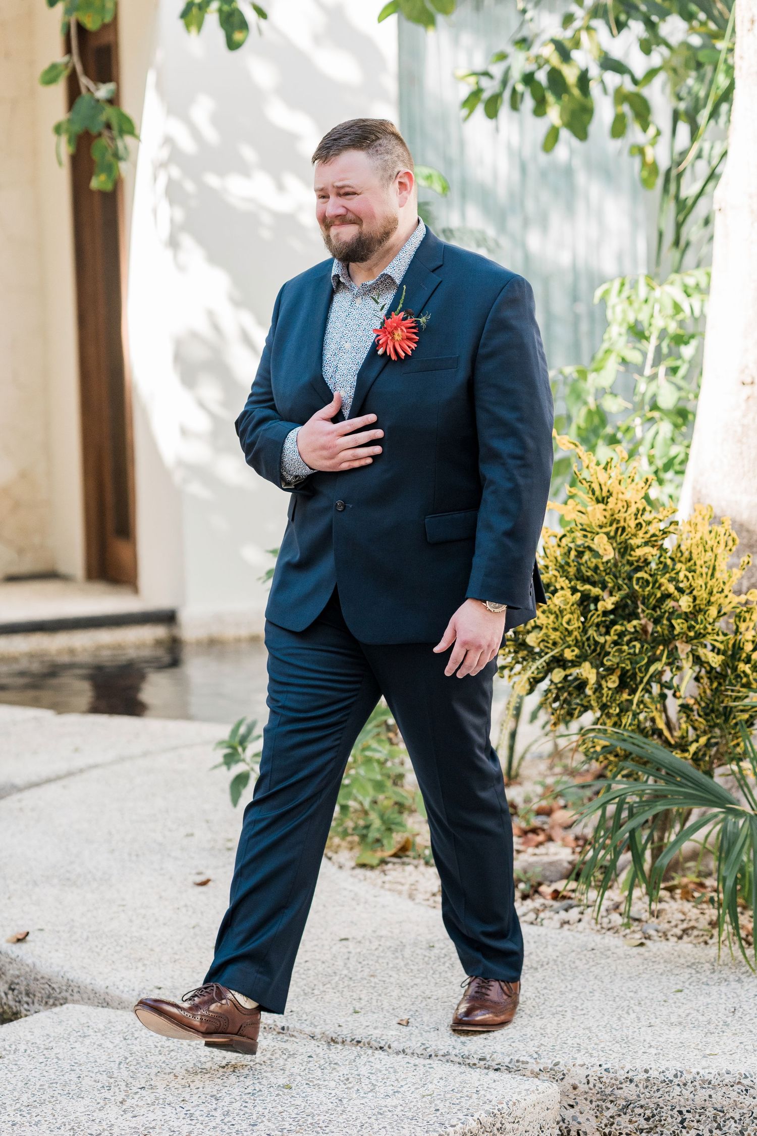 Secrets Akumal Wedding - Groom in navy blue suit with red boutonniere smiling outdoors near greenery on wedding day.