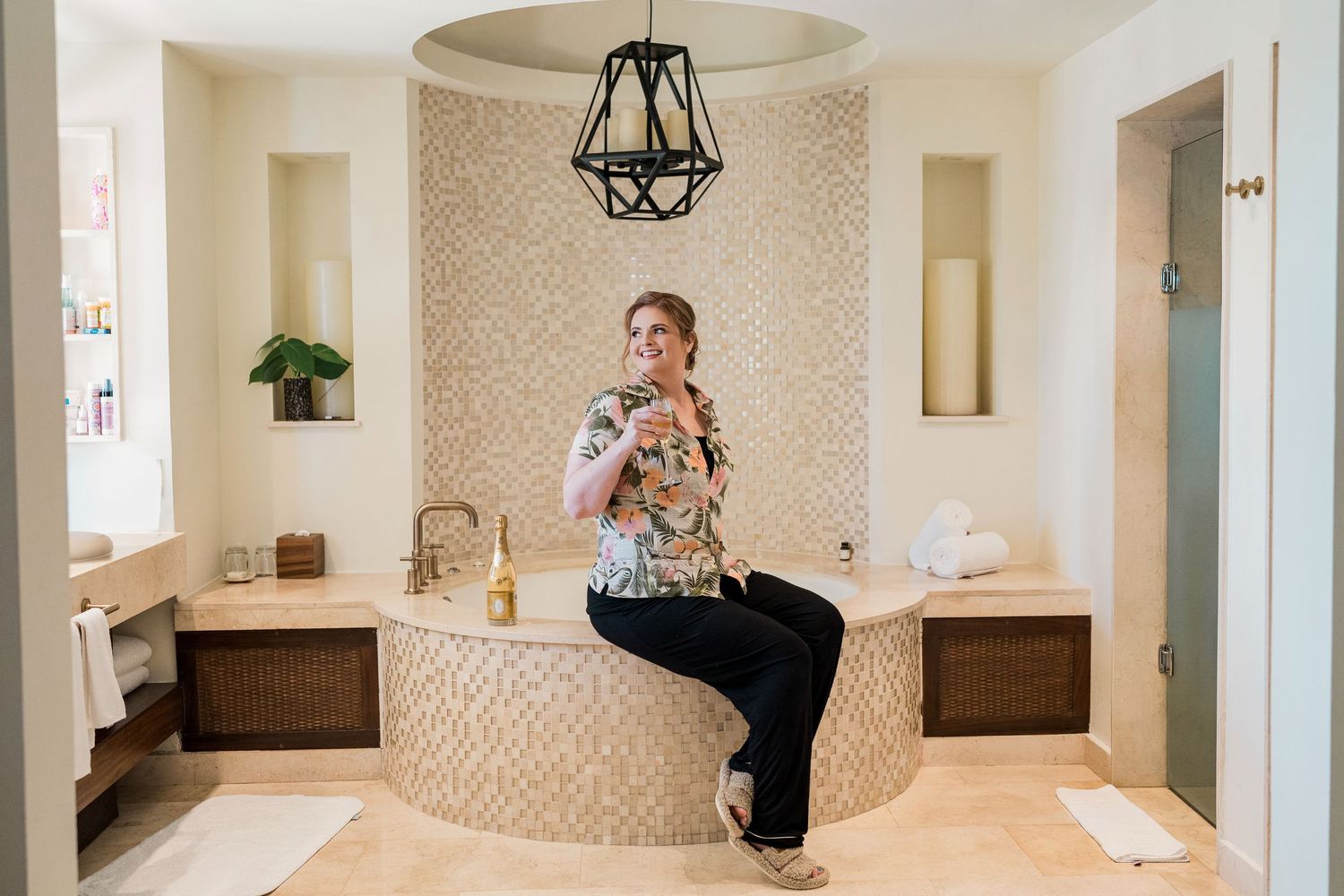 Woman sitting on edge of elegant freestanding bathtub in luxurious beige tiled bathroom with chandelier.
