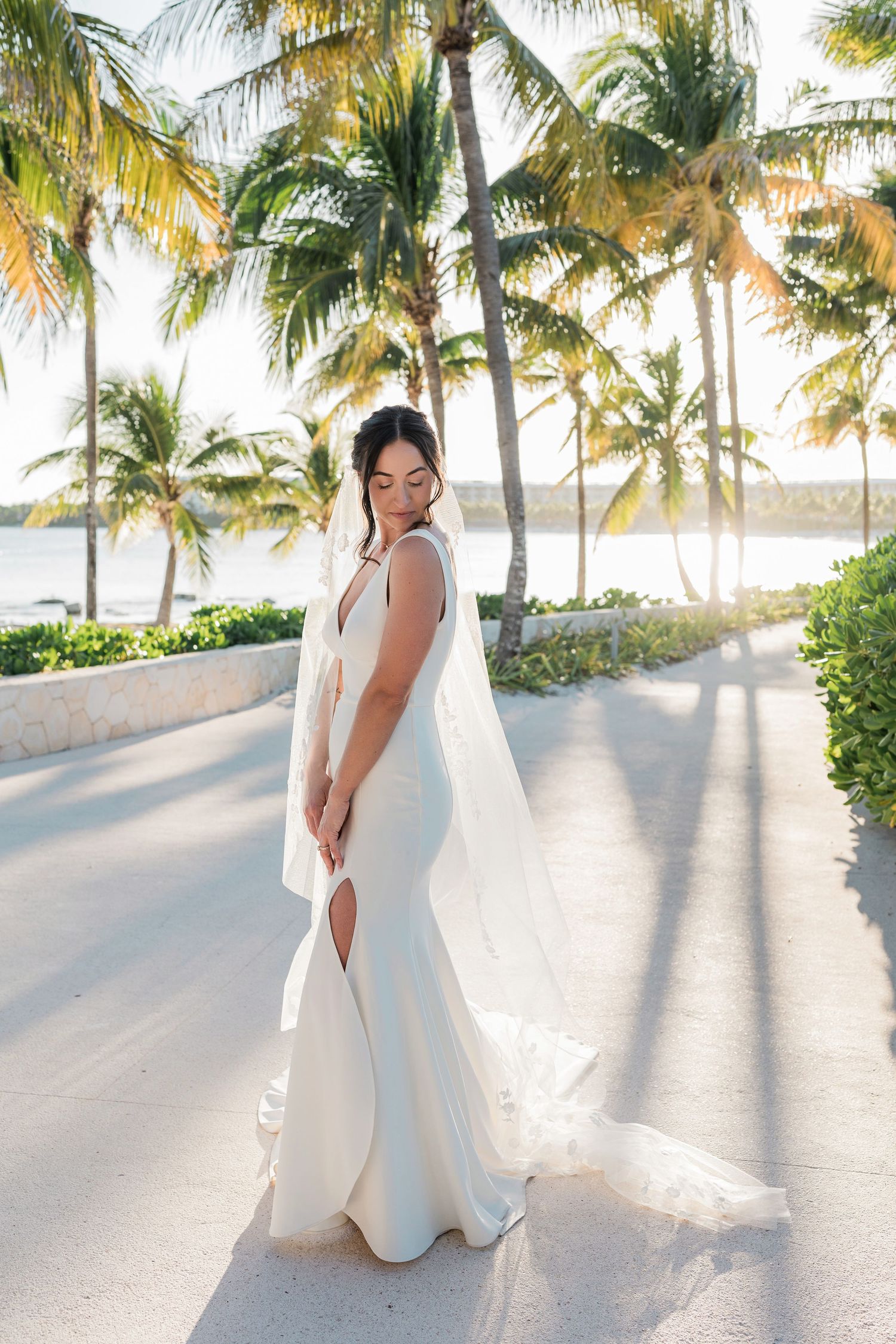 Bride in elegant white gown and veil poses among tropical palm trees near waterfront at golden hour.