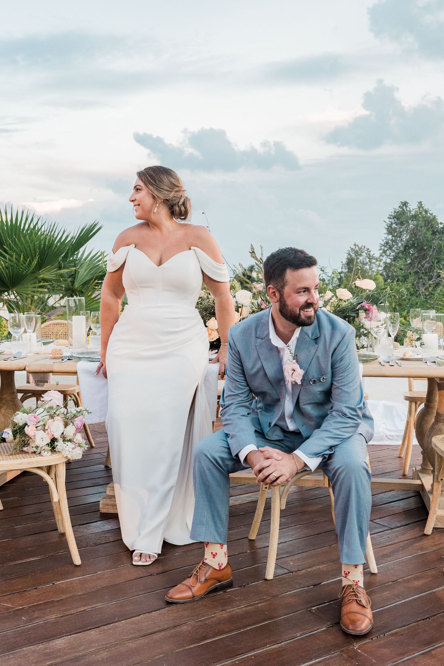 Bride in white off-shoulder gown stands beside seated groom in light blue suit at outdoor wedding reception on wooden deck.