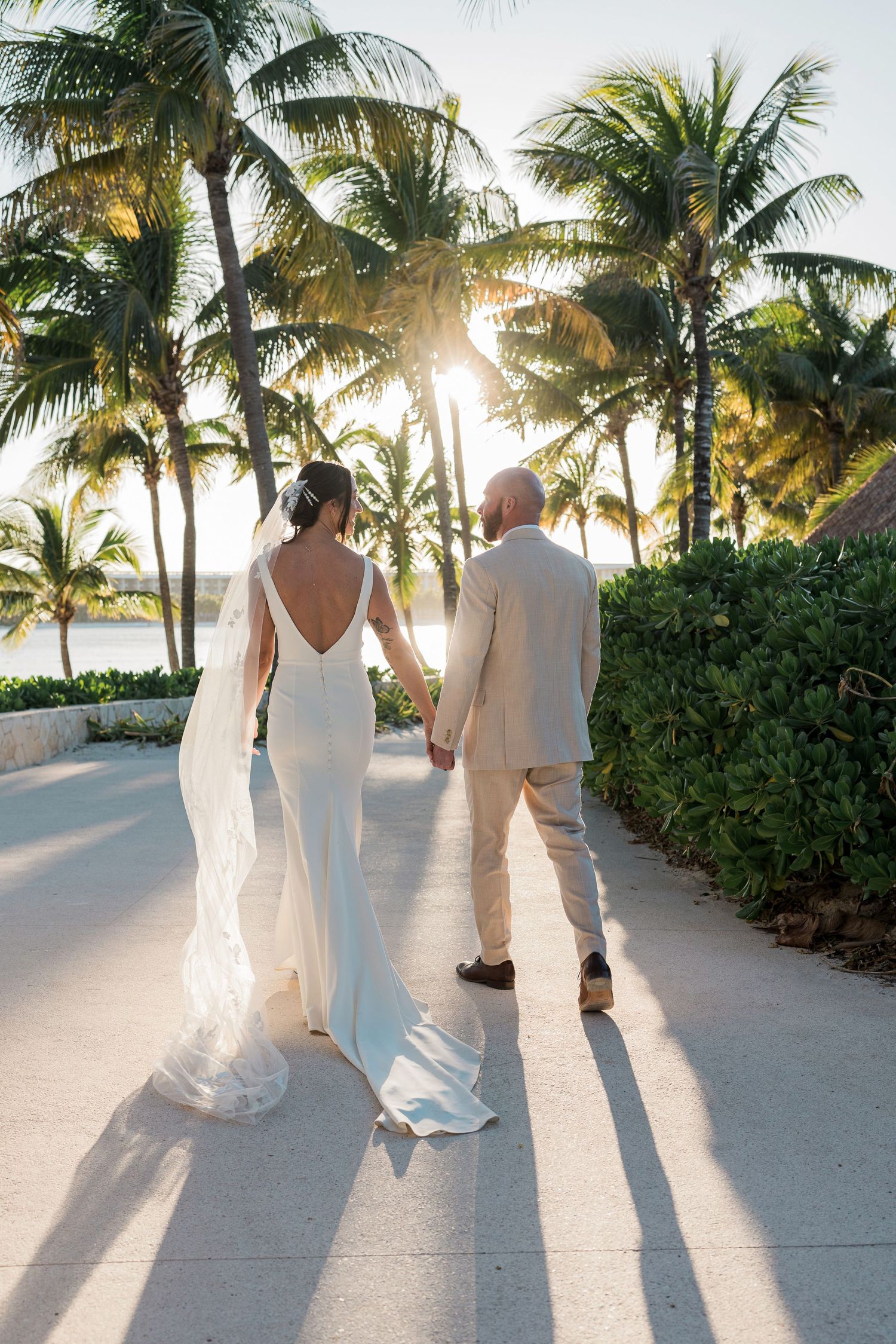 Bride and groom walk hand-in-hand along a tropical path lined with palm trees at sunset during their destination wedding.