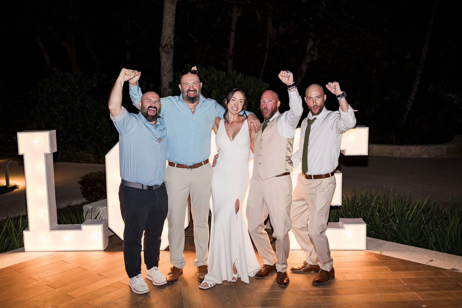 A joyful wedding group of five celebrating outdoors at night, with the bride in white and guests cheering.