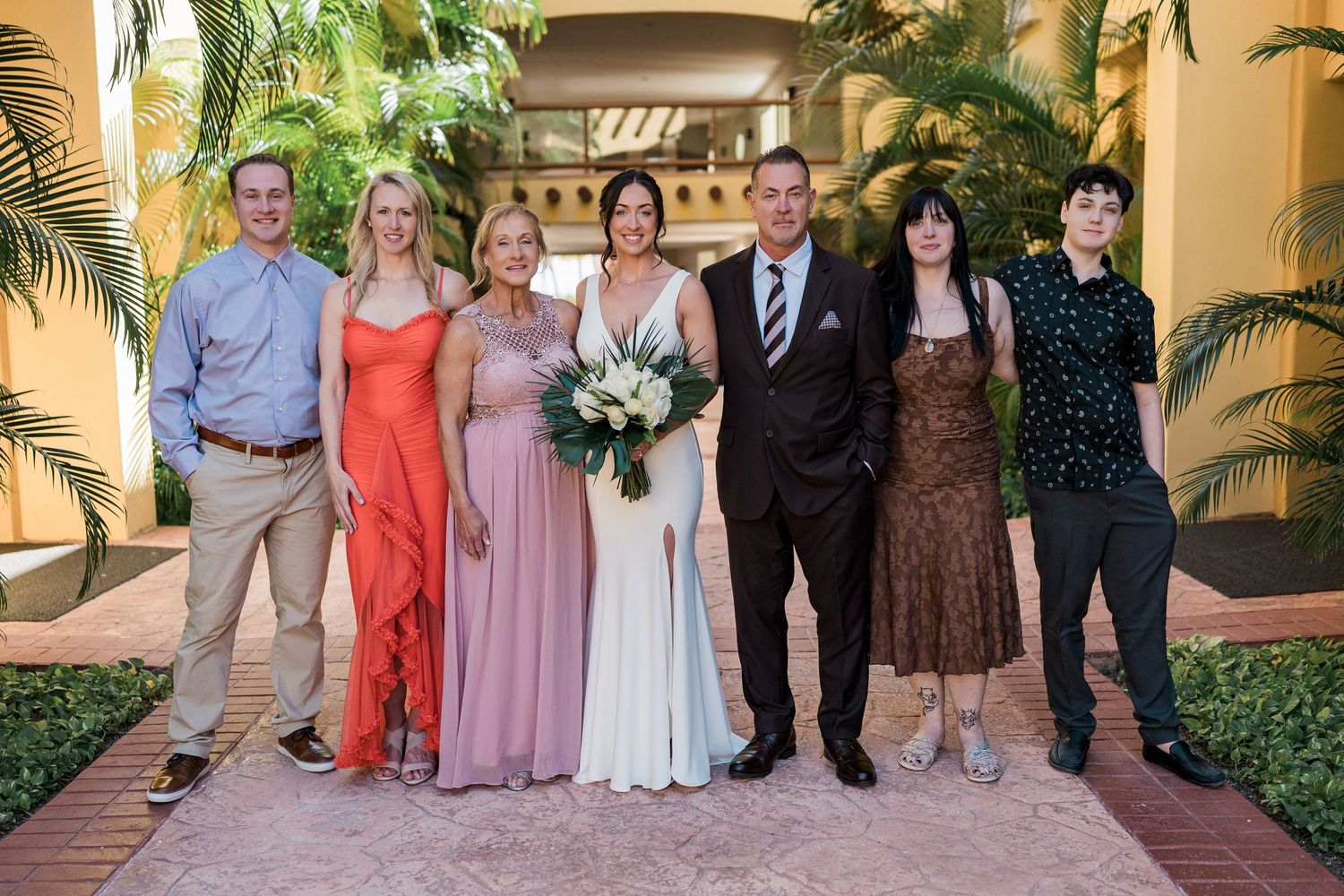 Wedding party of six posing together outdoors at elegant venue with tropical plants, bride in white gown holding bouquet.