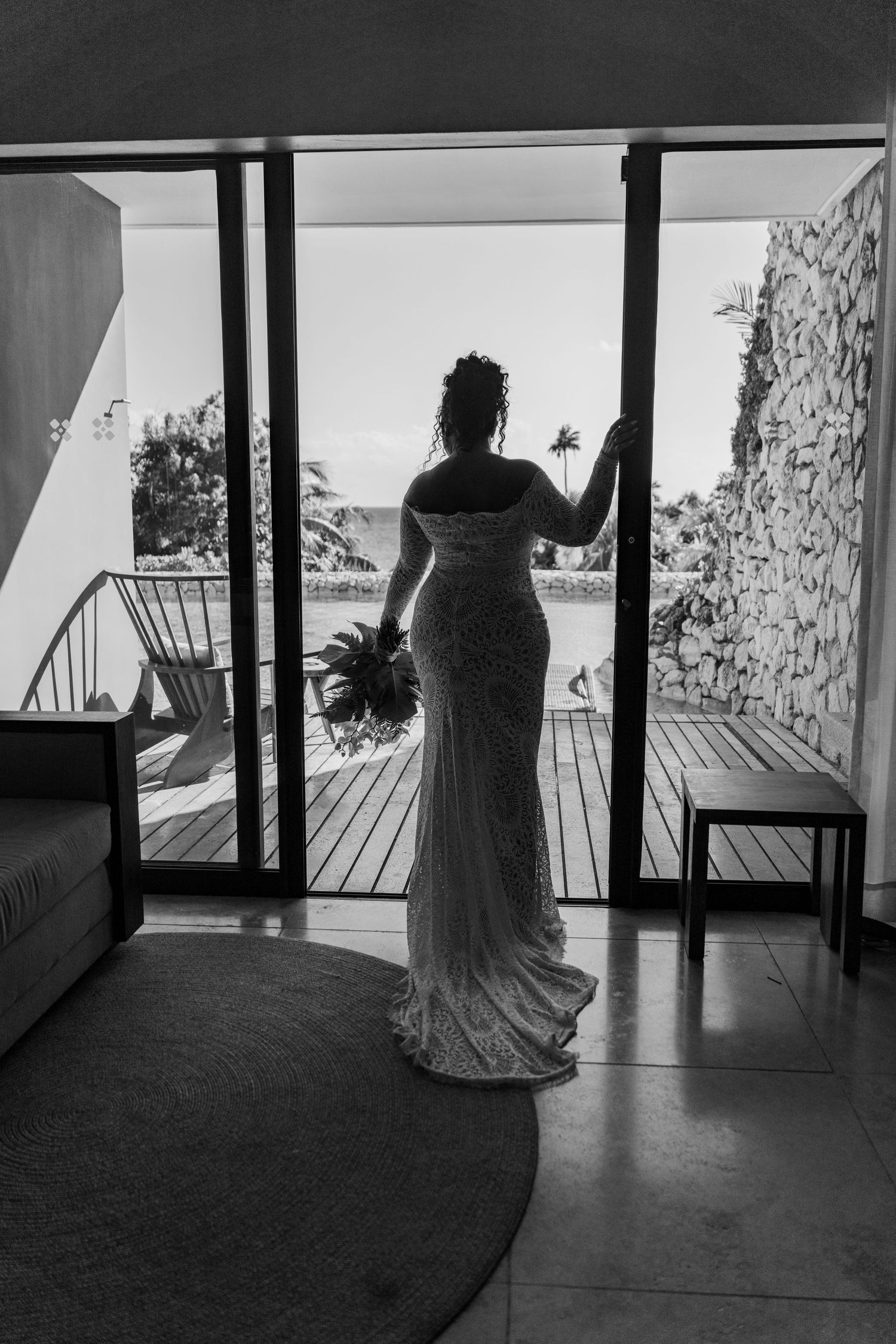 Xcaret Arte Wedding - Black and white photo of a bride in a lace gown standing at open glass doors overlooking a tropical resort balcony.