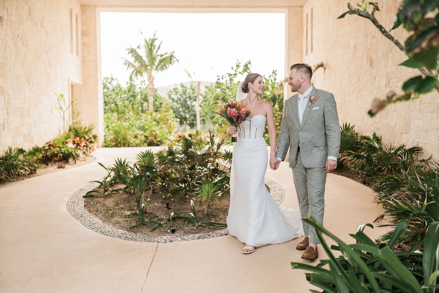 Bride and groom hold hands in courtyard garden with tropical plants and archway.