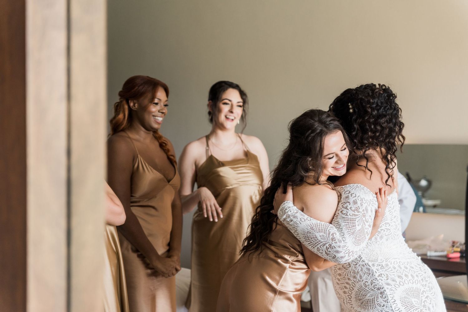 Xcaret Arte Wedding - Bride hugging bridesmaid in lace gown while two bridesmaids in gold satin dresses smile during wedding preparations.