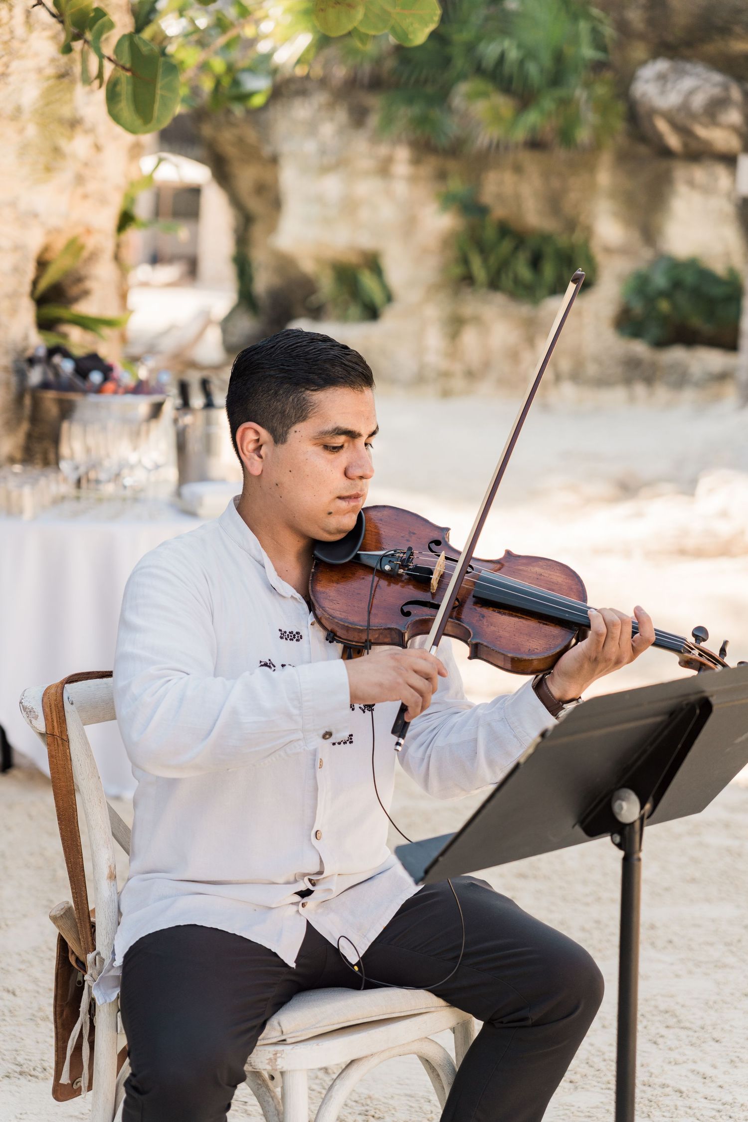 Xcaret Arte Wedding - Young man in white shirt plays violin while seated outdoors at an elegant event with music stand.