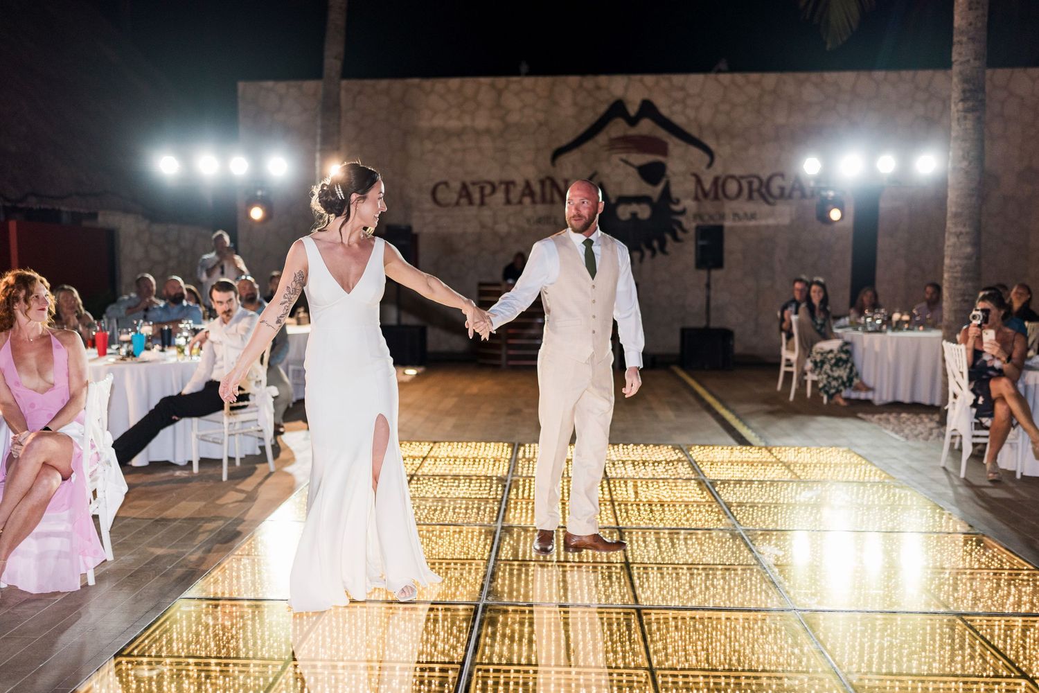 Bride and groom hold hands on a glowing gold dance floor at an outdoor wedding reception under Captain Morgan signage.