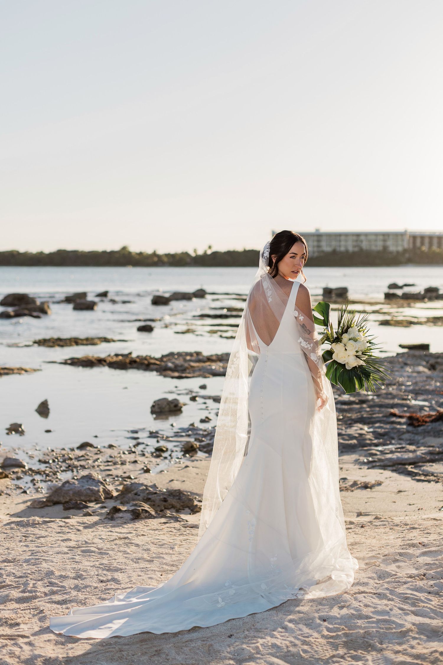 Bride in white lace gown with veil holds bouquet on rocky beach at sunset.