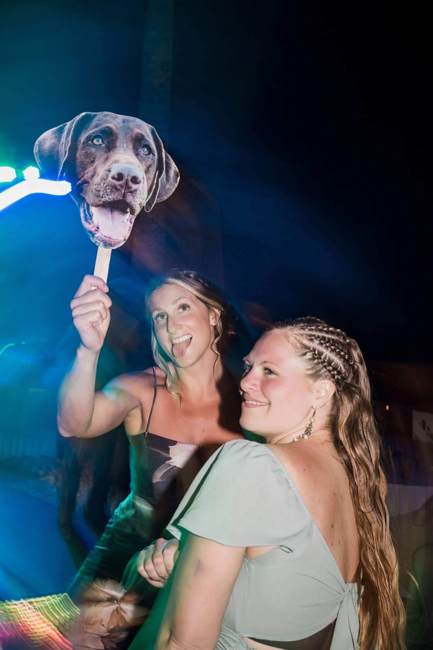 Two women smiling at a party, one holding a dog face prop on a stick under colorful lights.