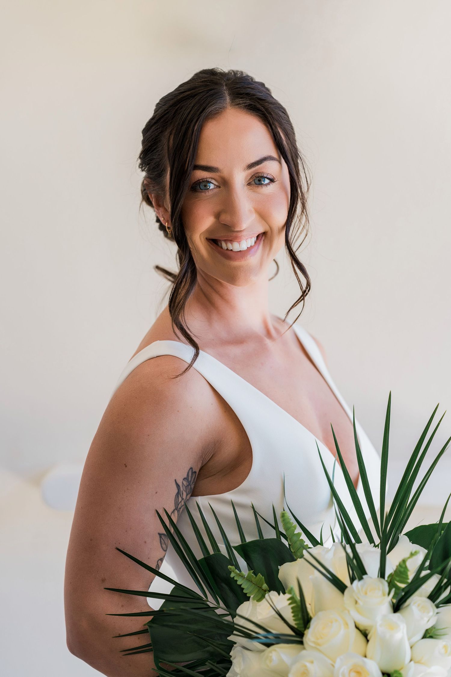 Smiling bride in white dress holding white floral bouquet with tropical leaves, showcasing elegant updo and arm tattoo.