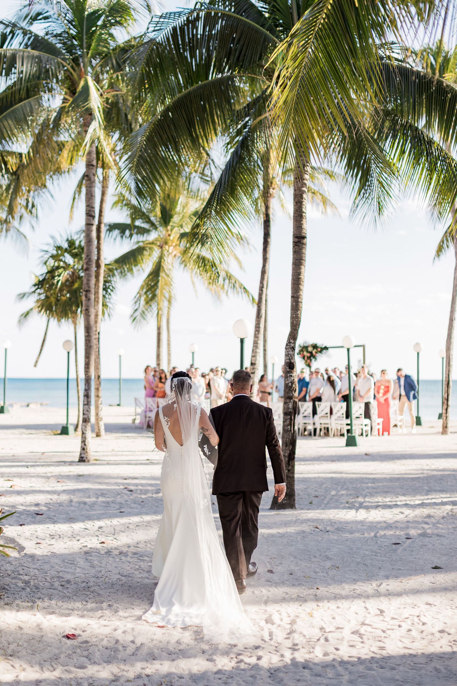 Bride and groom walk hand-in-hand toward a beach wedding ceremony surrounded by tall palm trees on white sand.