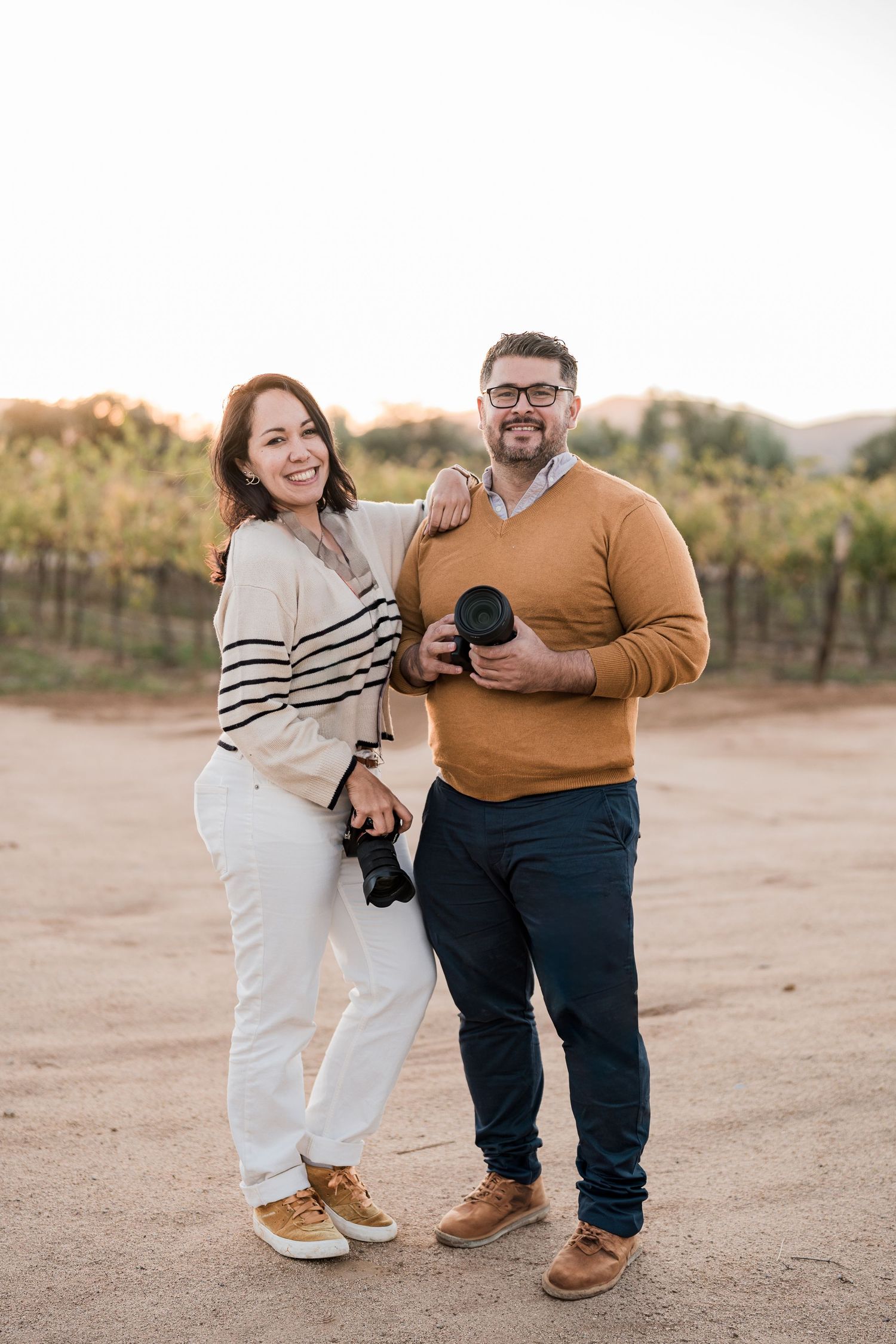 A smiling couple holding cameras stands together in a sunny vineyard, dressed in casual attire.
