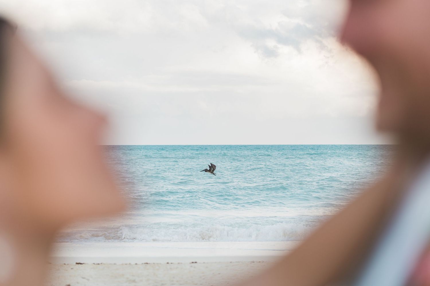 A bird flies over turquoise ocean waves framed by two blurred figures on a sandy beach.