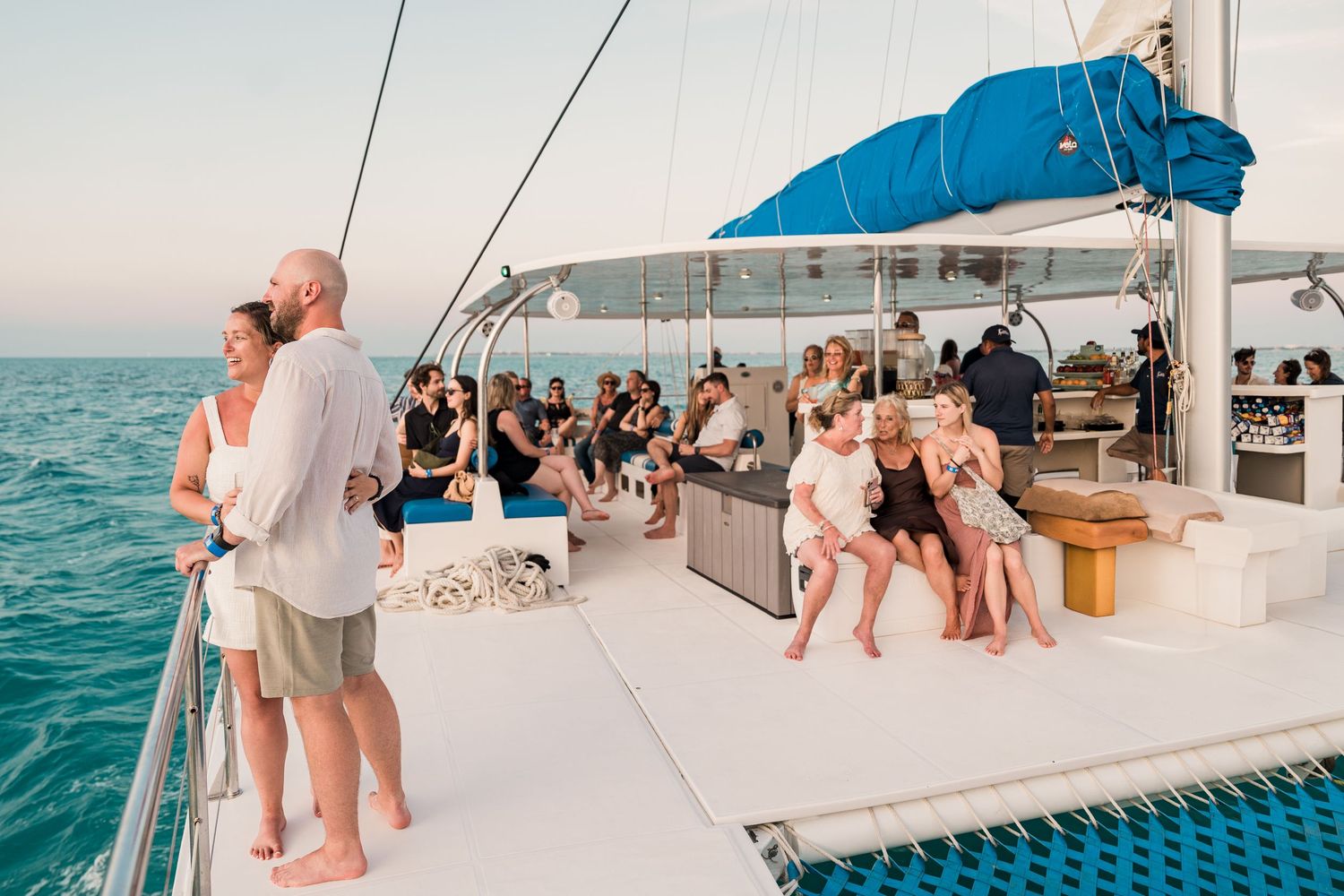 Guests socializing on a luxury catamaran deck at sunset, with calm turquoise ocean waters in the background.