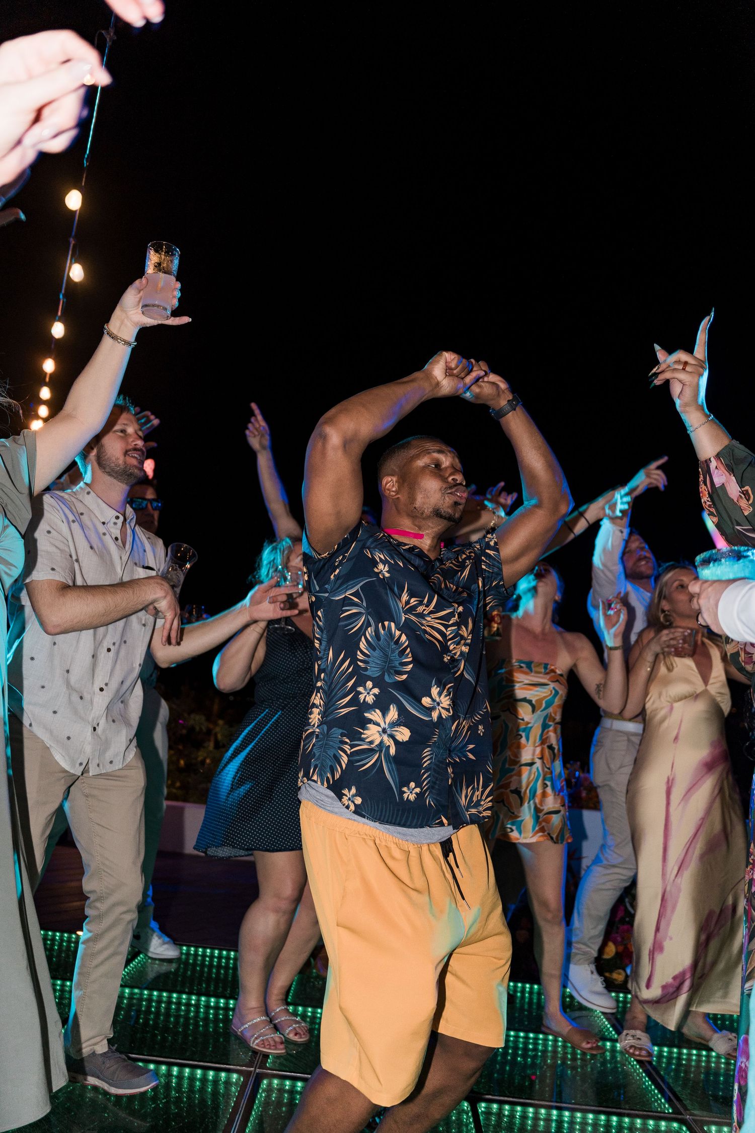 Diverse group of people dancing at an outdoor night party, with a man in floral shirt and yellow shorts in the foreground.