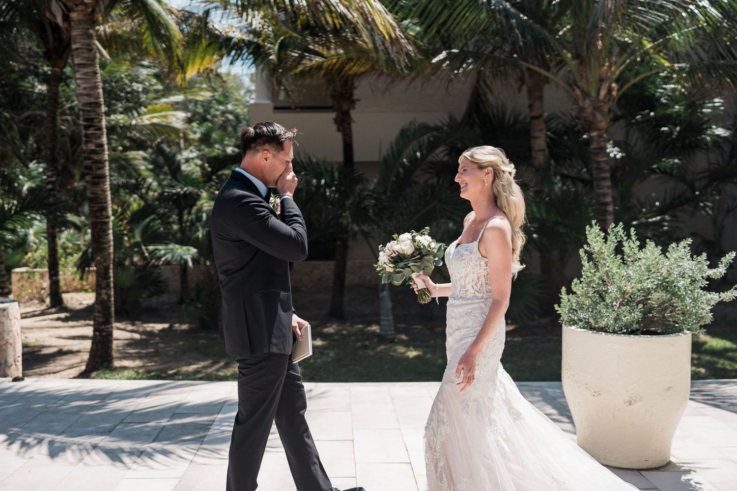 Groom emotional at first look seeing bride in white lace wedding gown holding bouquet outdoors with palm trees.